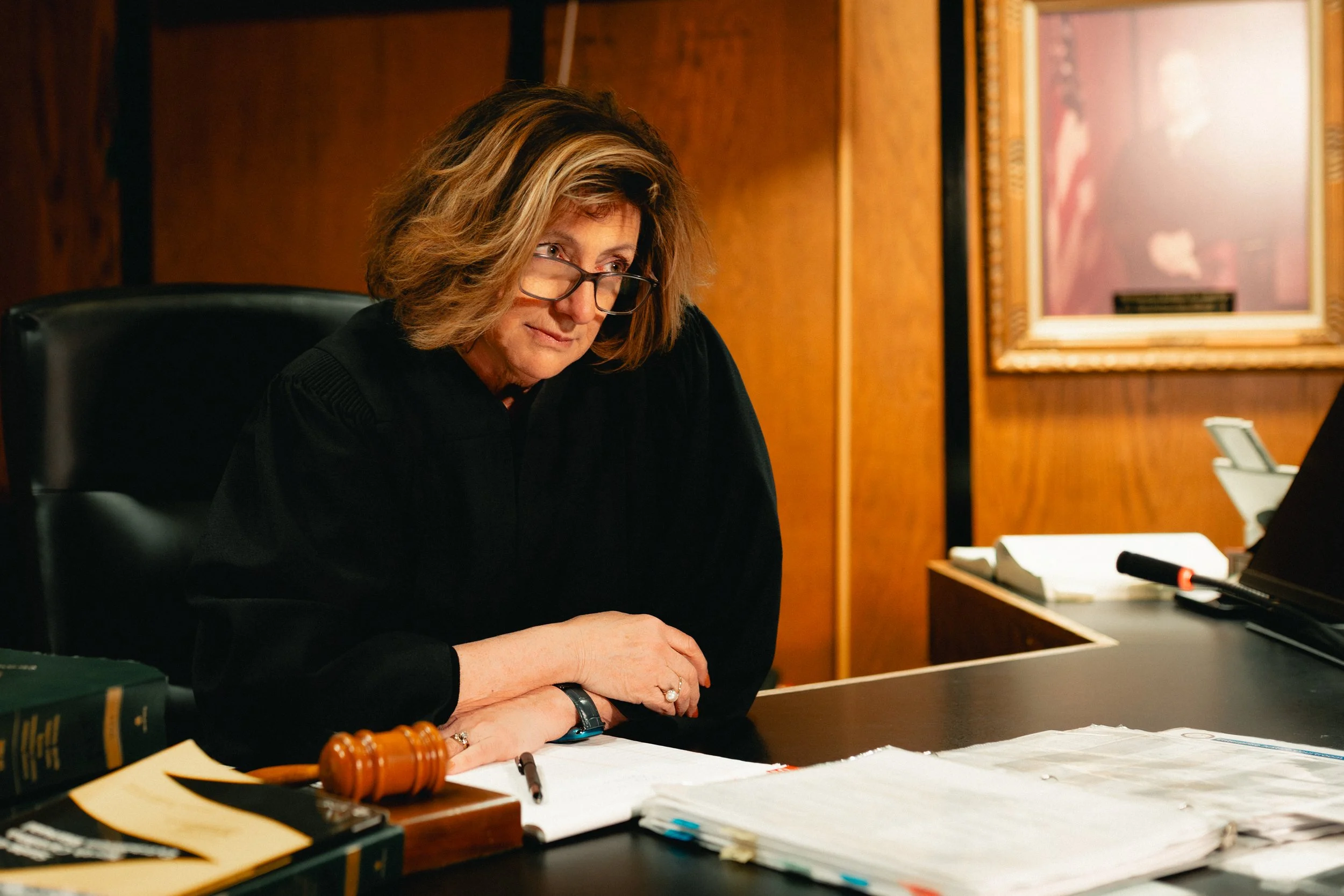 A woman judge in black robes sitting at a court desk, looking down thoughtfully with glasses on, surrounded by legal papers, a gavel, and office equipment in a wood-paneled courtroom.