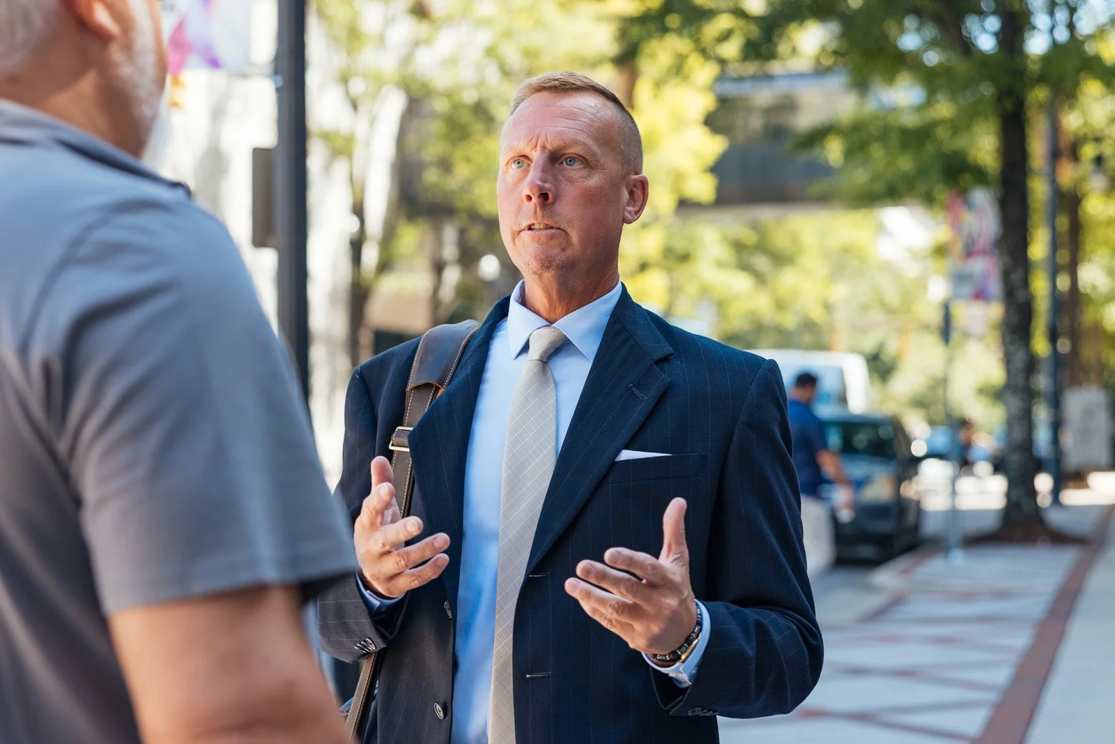A man in a navy suit and light gray tie talking to another man in a gray T-shirt on a city sidewalk with trees and parked cars in the background.