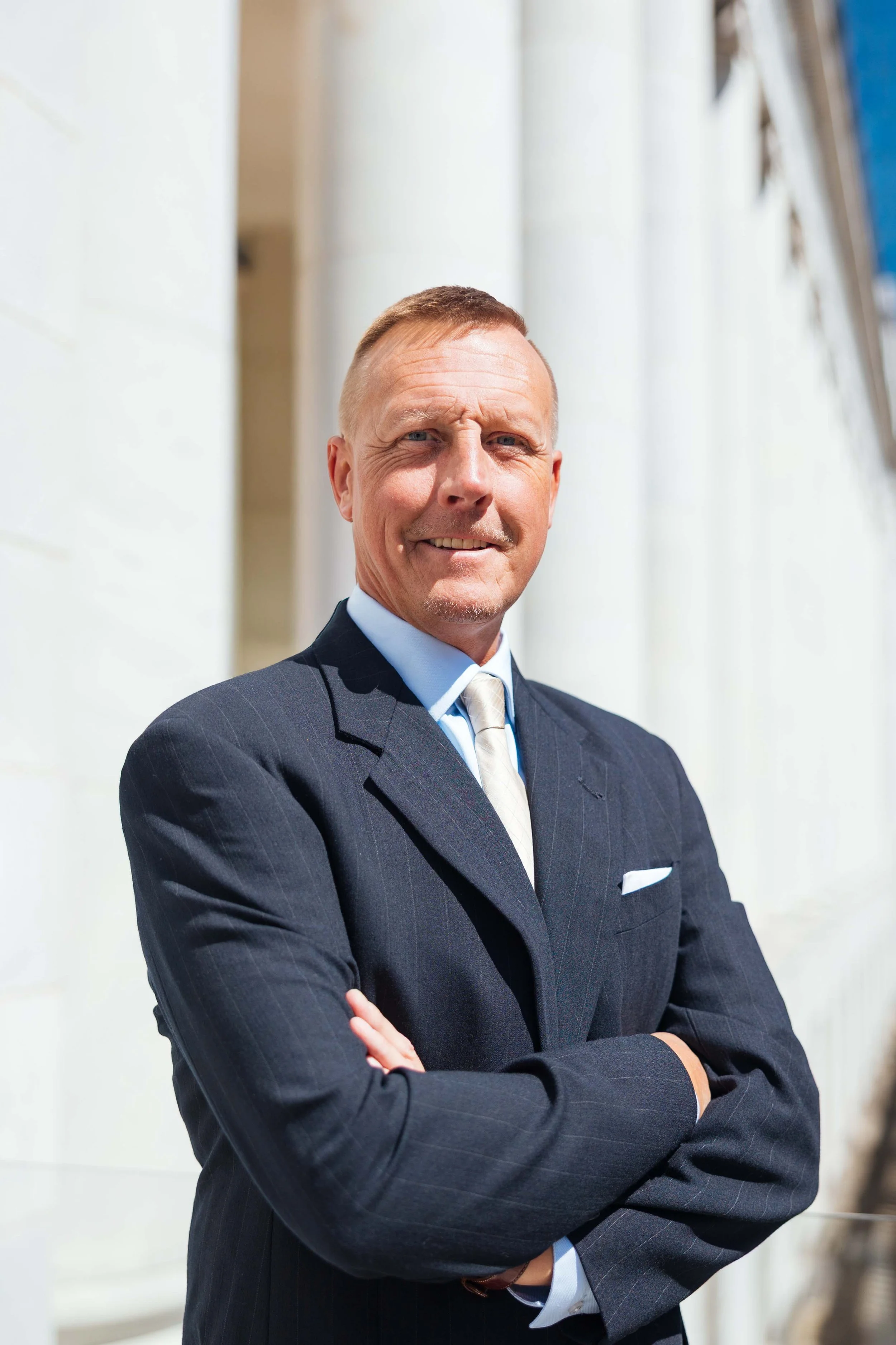 A middle-aged man in a dark business suit with a white shirt and beige tie standing outdoors with crossed arms, smiling, in front of white columns.