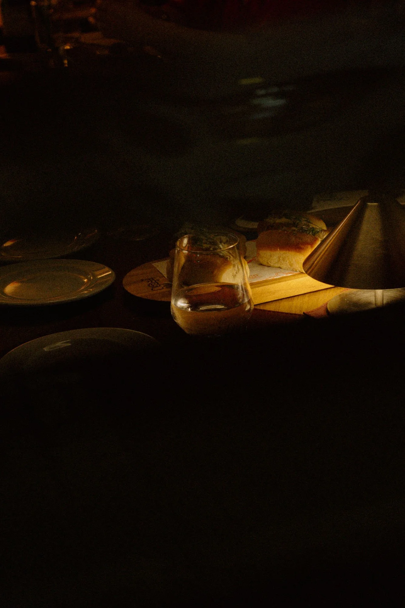 Imagen de una comida en un entorno oscuro, con un plato plano, un vaso con agua y una tabla de madera con un sándwich, acompañados de una lámpara de pie.
