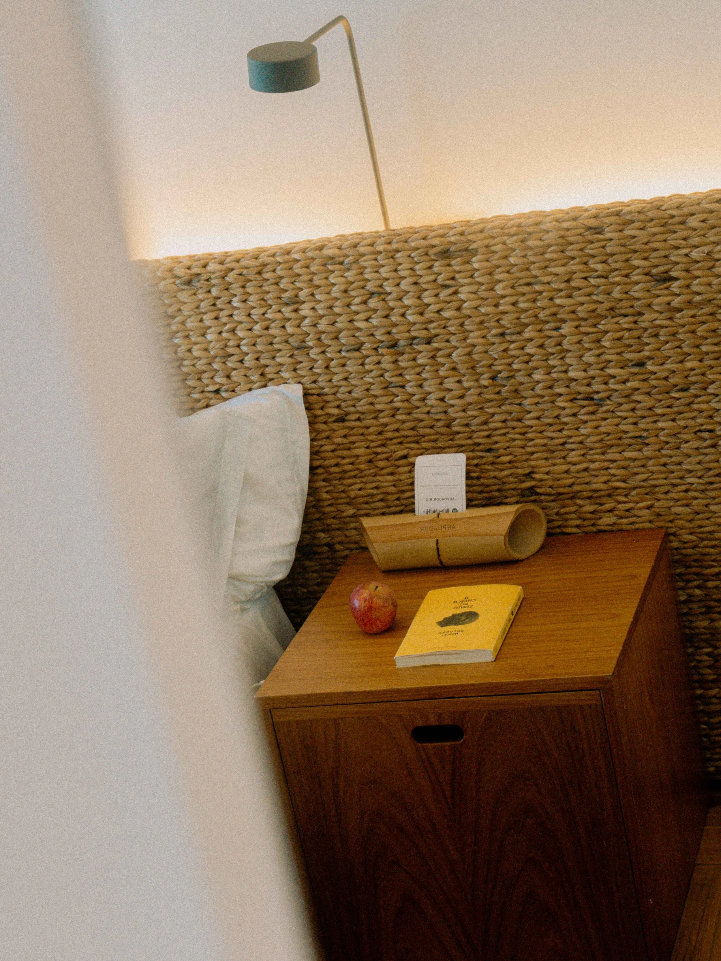 Una cama con cabecera de ratán, una almohada blanca, una lámpara de pared moderna, una libreta amarilla, una manzana roja, una caja de cerillas amarilla, y una mesita de madera.