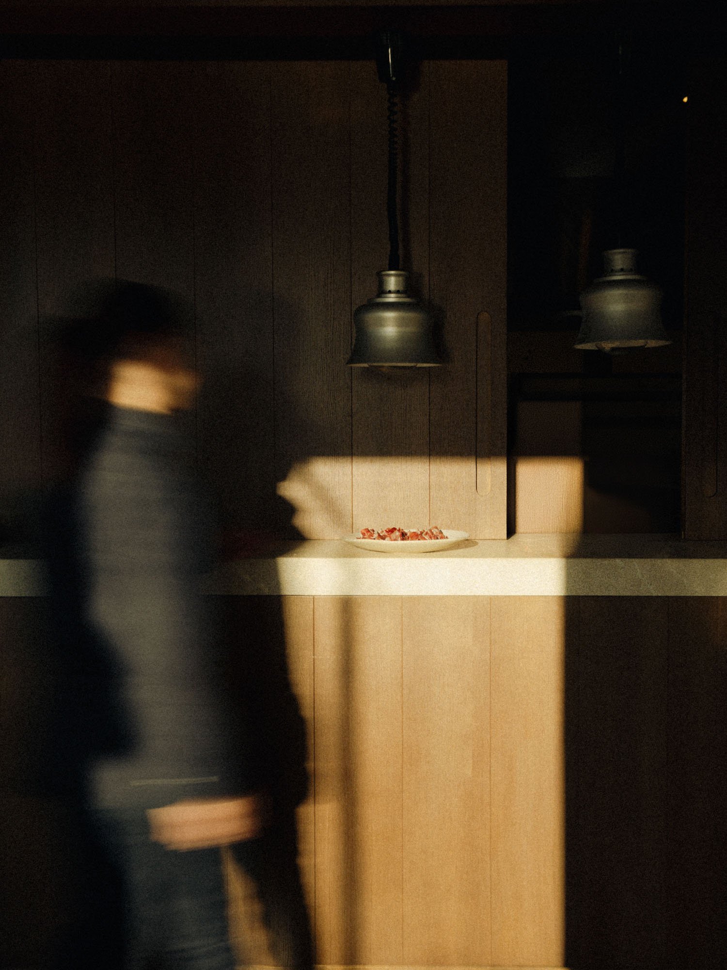 Imagen de una oficina moderna con paredes de madera oscura y lámparas colgantes de metal. En primer plano, una persona borrosa pasa frente a la cámara mientras una bandeja con trozos de carne se encuentra sobre una superficie en el centro.