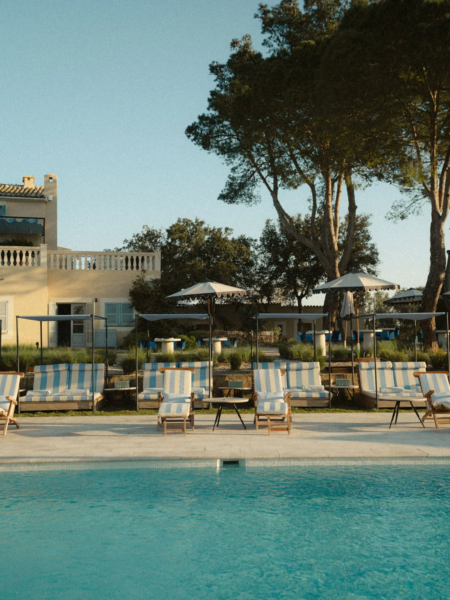 Piscina con sillas y sombrillas blancas en un área al aire libre, árboles y un edificio en el fondo, cielo despejado.
