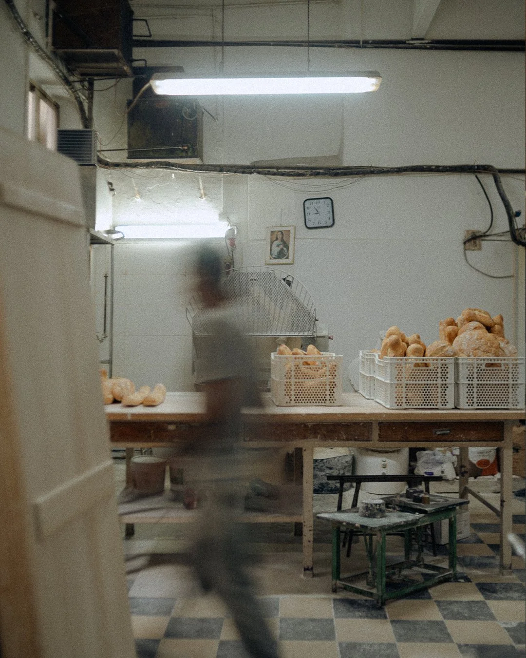 Baker moving through the kitchen of Horno El Reloj, a traditional bakery in El Cabanyal, Valencia, with fresh bread in baskets on the worktable