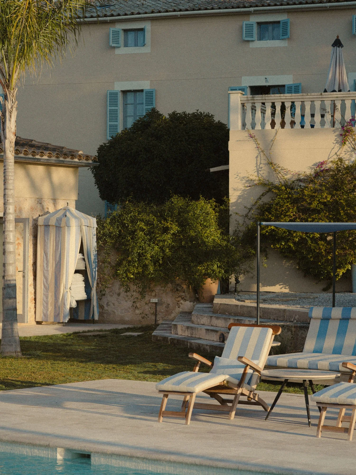 Áreas de una piscina y zona de descanso con sillas y sombrillas, cerca de una casa con ventanas con contraventanas azules.