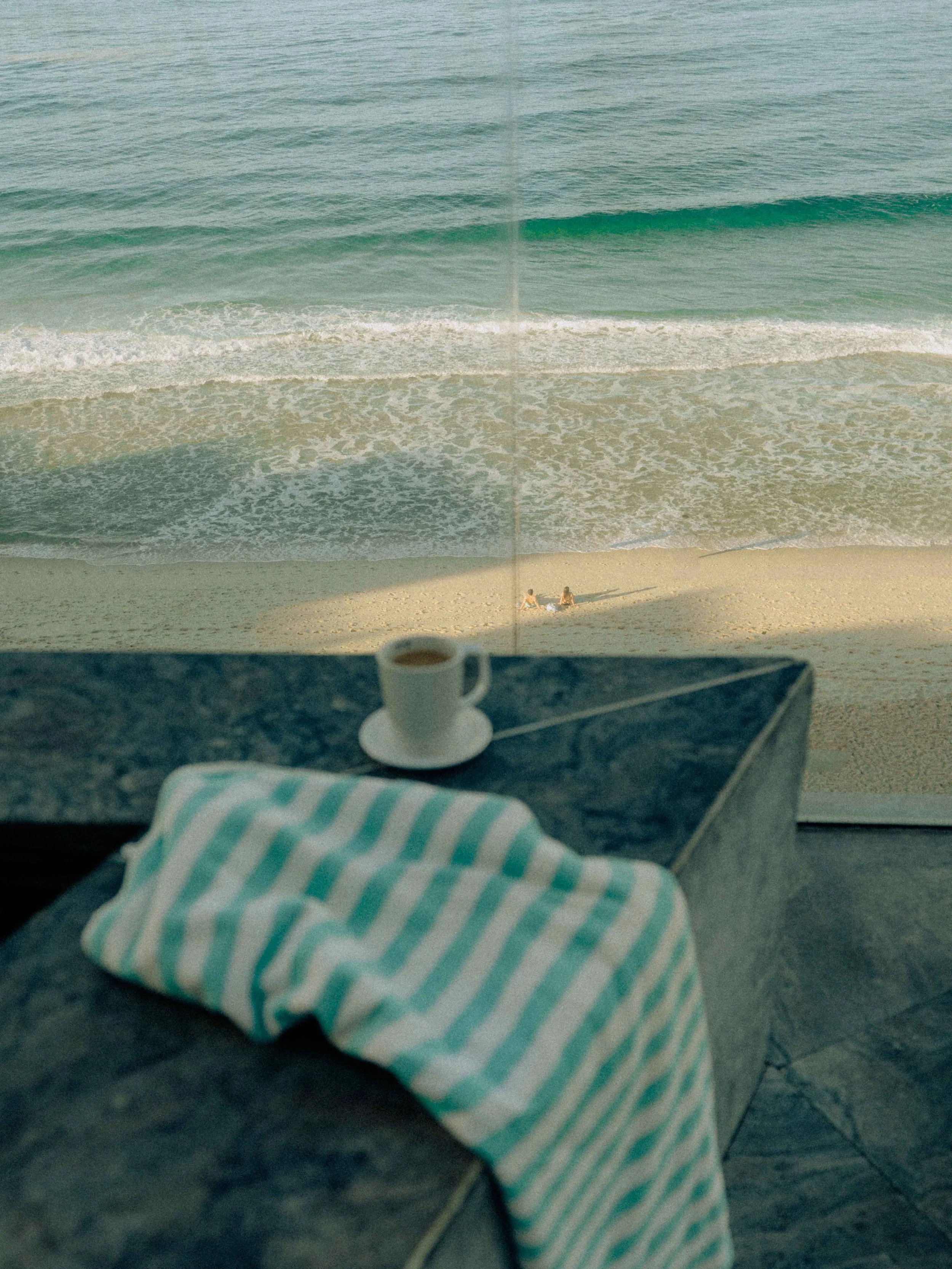 Vista de una playa con olas rompiendo en la orilla, dos personas sentadas en la arena, vista desde una mesa con una taza blanca y un paño azul y blanco en primer plano.