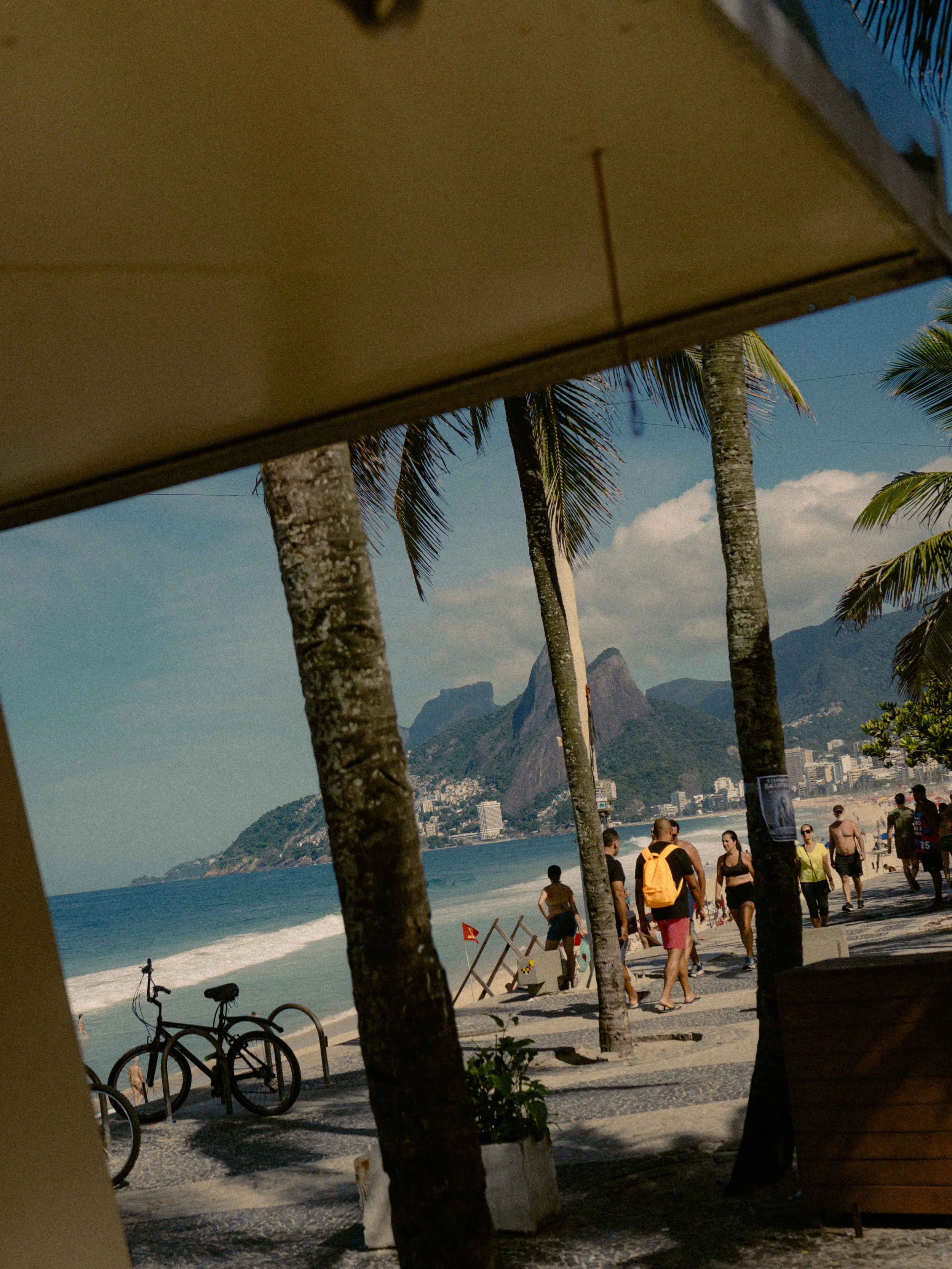 Vista de la playa con palmeras, personas caminando, bicicletas estacionadas y el mar, con montañas al fondo, vista desde un toldo en un día soleado.