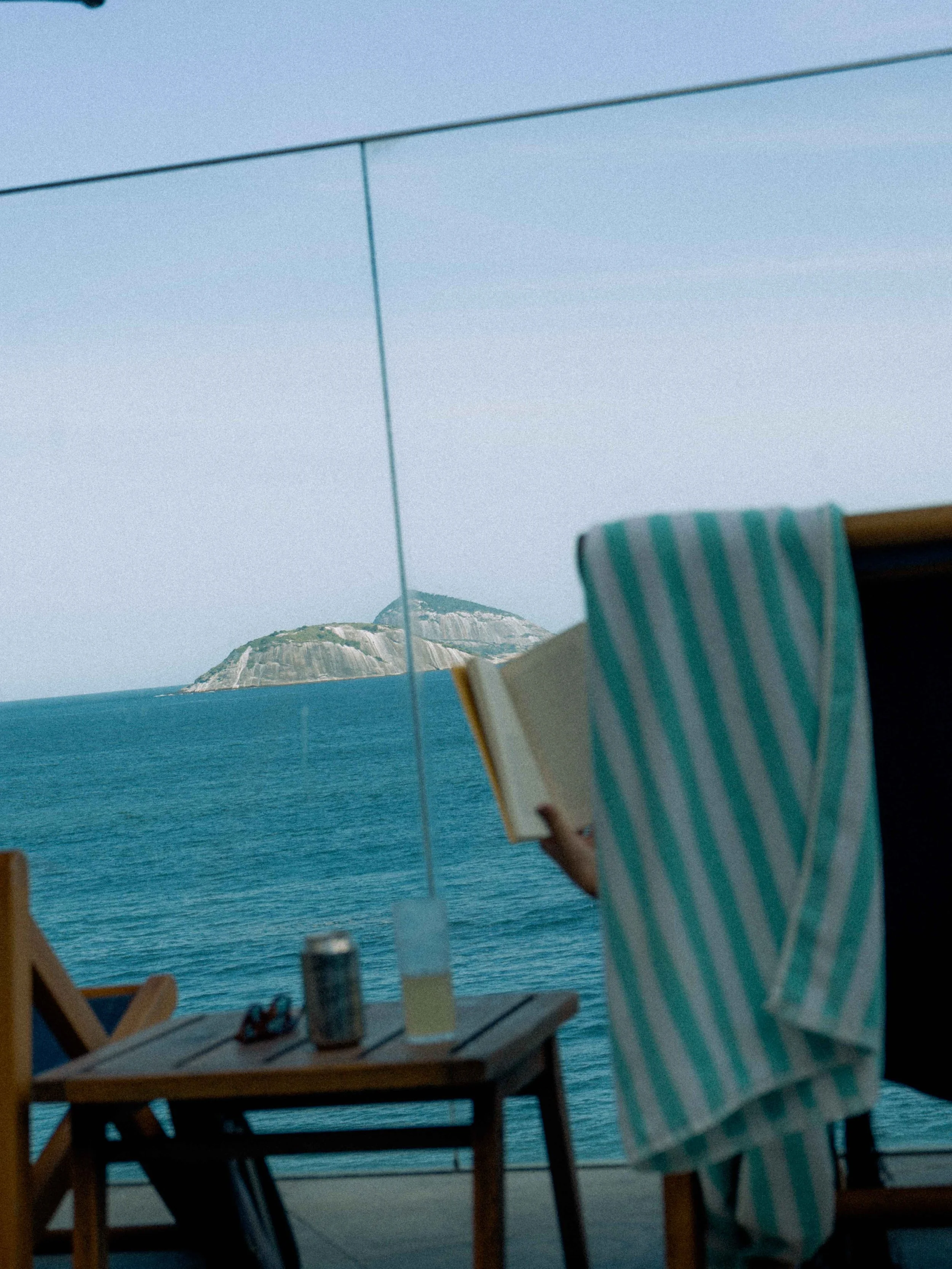 Persona leyendo un libro en un balcón con vista al mar y una montaña, con una mesa y una silla de madera, y una camisa a rayas