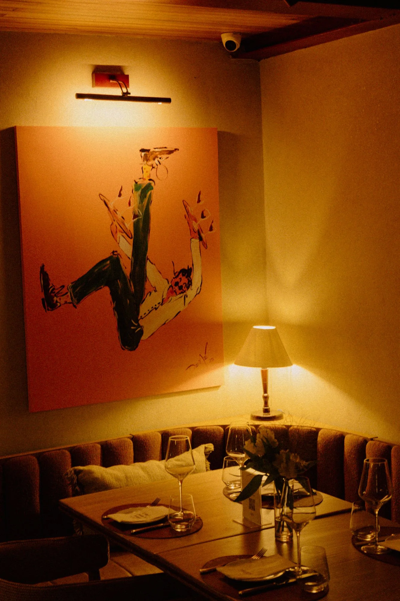 Una sala de comedor con una mesa decorada con copas y platos, un sofá morado y una lámpara en una mesa auxiliar, con un mural colorido de un hombre en la pared y una cámara de seguridad en el techo.