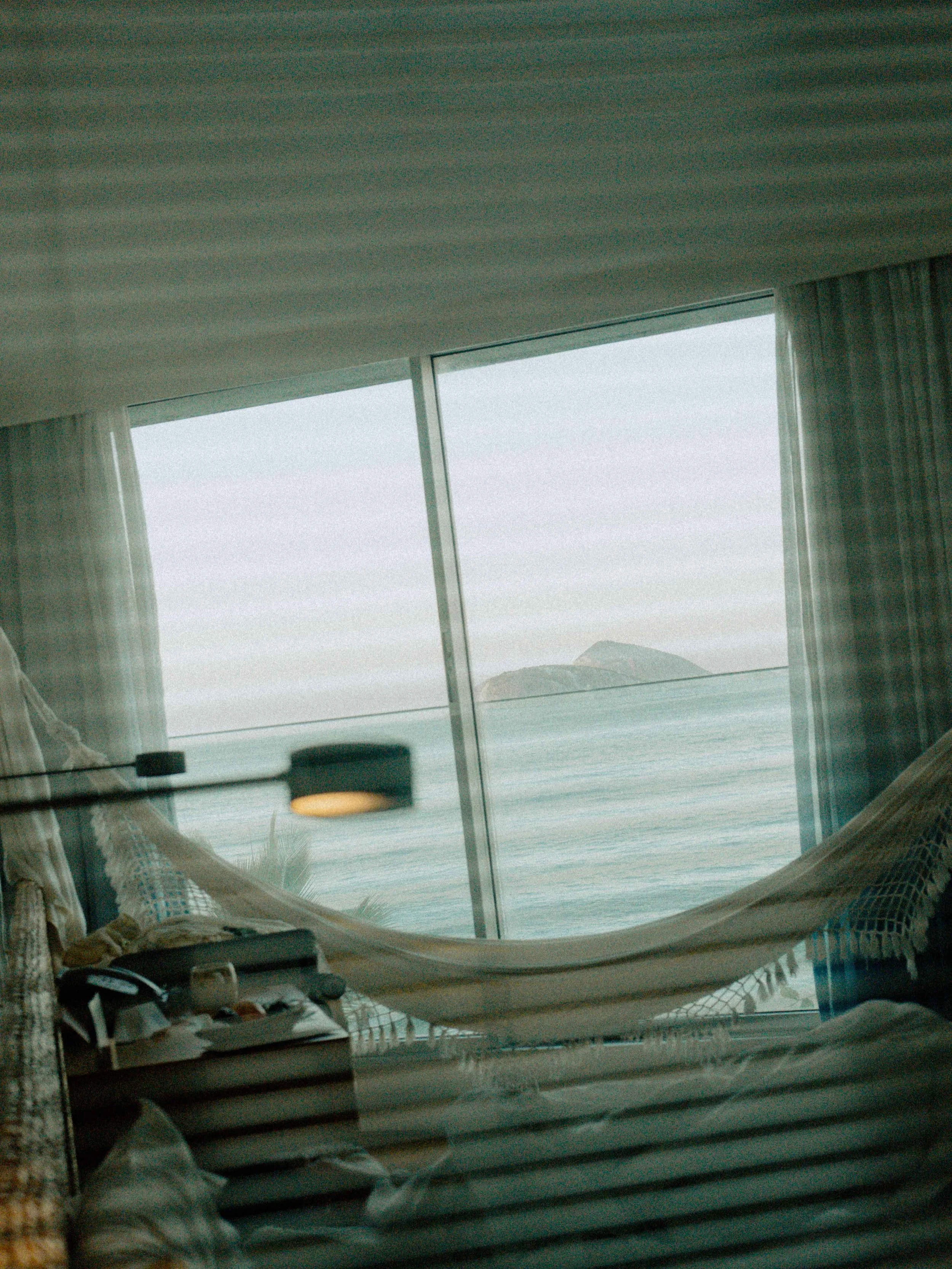 Vista desde una ventana grande con cortinas blancas, mostrando un paisaje de mar y montañas en la distancia, en un interior con muebles y objetos personales.