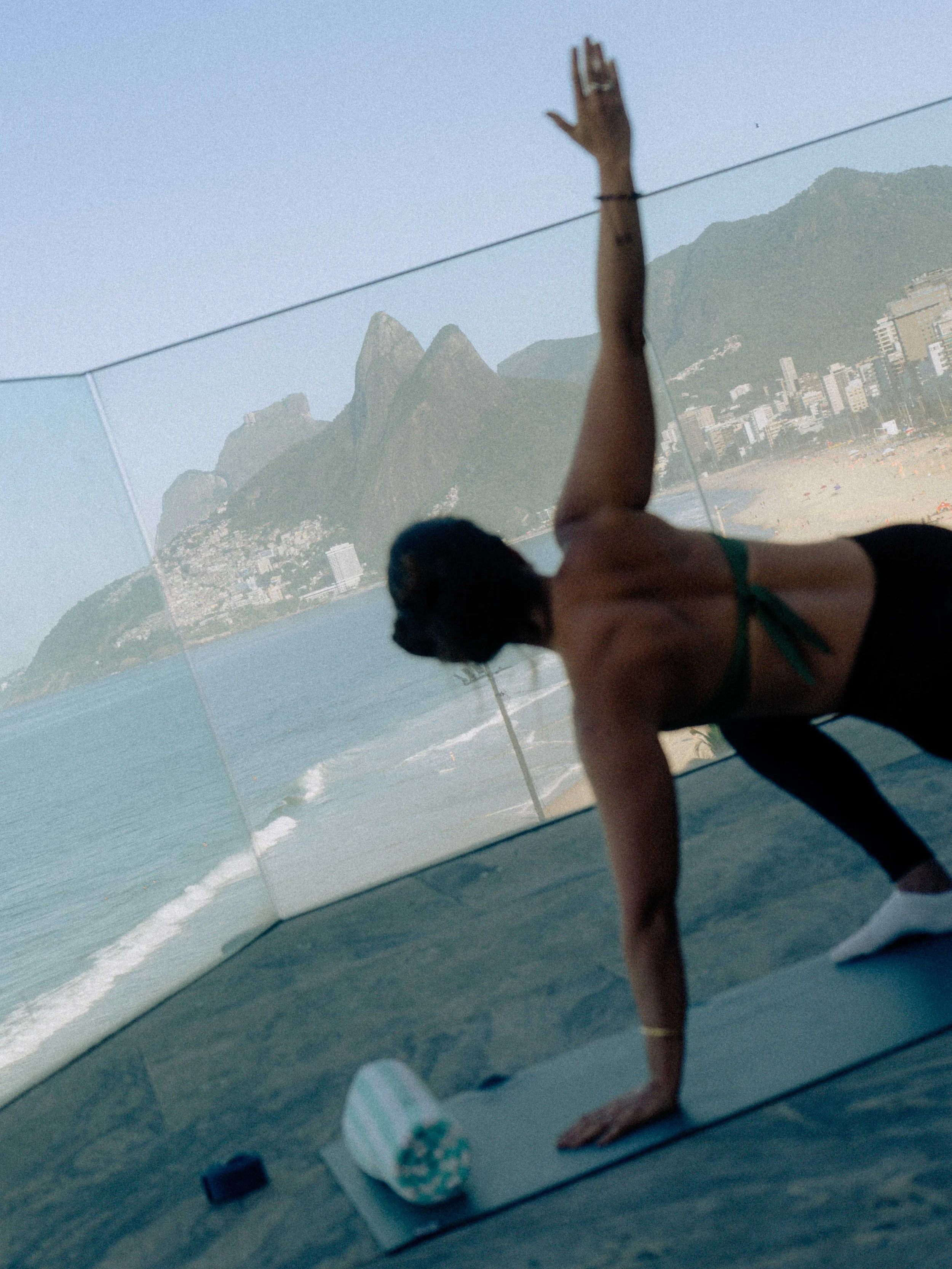 Reflejo en un espejo que muestra a una mujer haciendo yoga en una playa con montañas y edificios al fondo, mientras que ella se encuentra en el suelo en una postura de yoga.