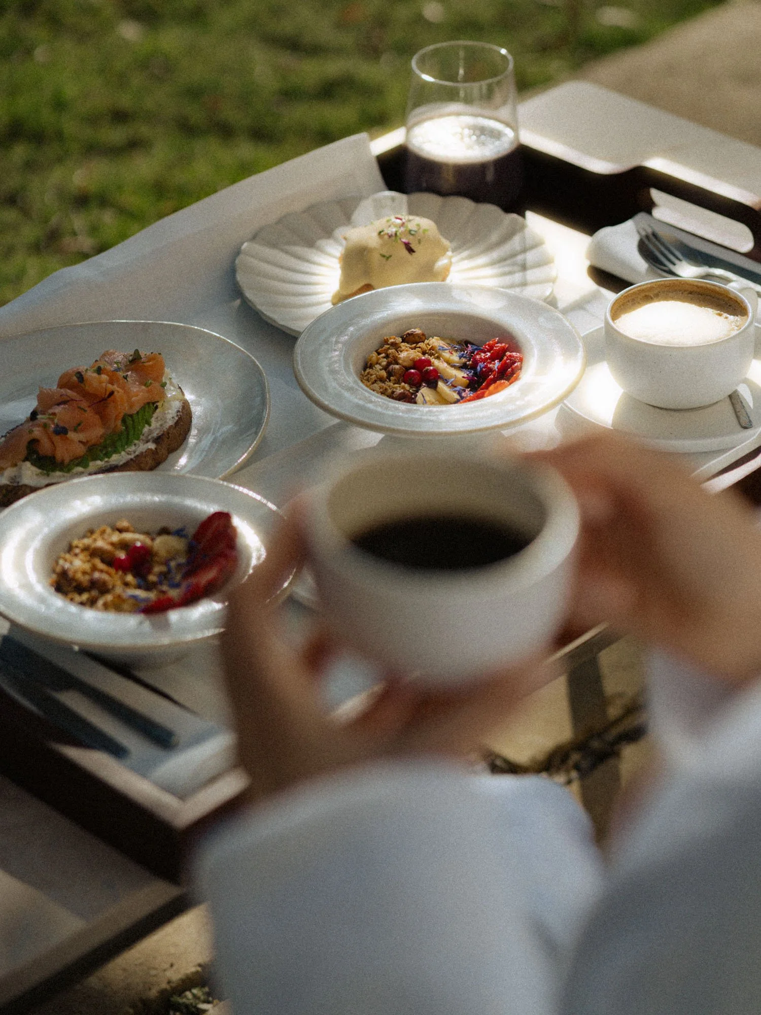 Mesa con comida y bebidas, incluyendo un vaso con líquido, una taza con café, y diferentes platos con alimentos, en un entorno al aire libre.