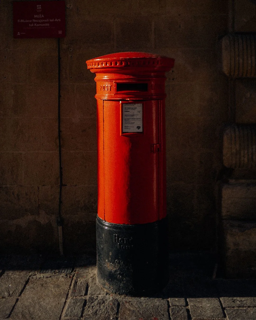 A red British postal mailbox standing on a cobblestone sidewalk at night, with a dimly lit brick wall in the background.
