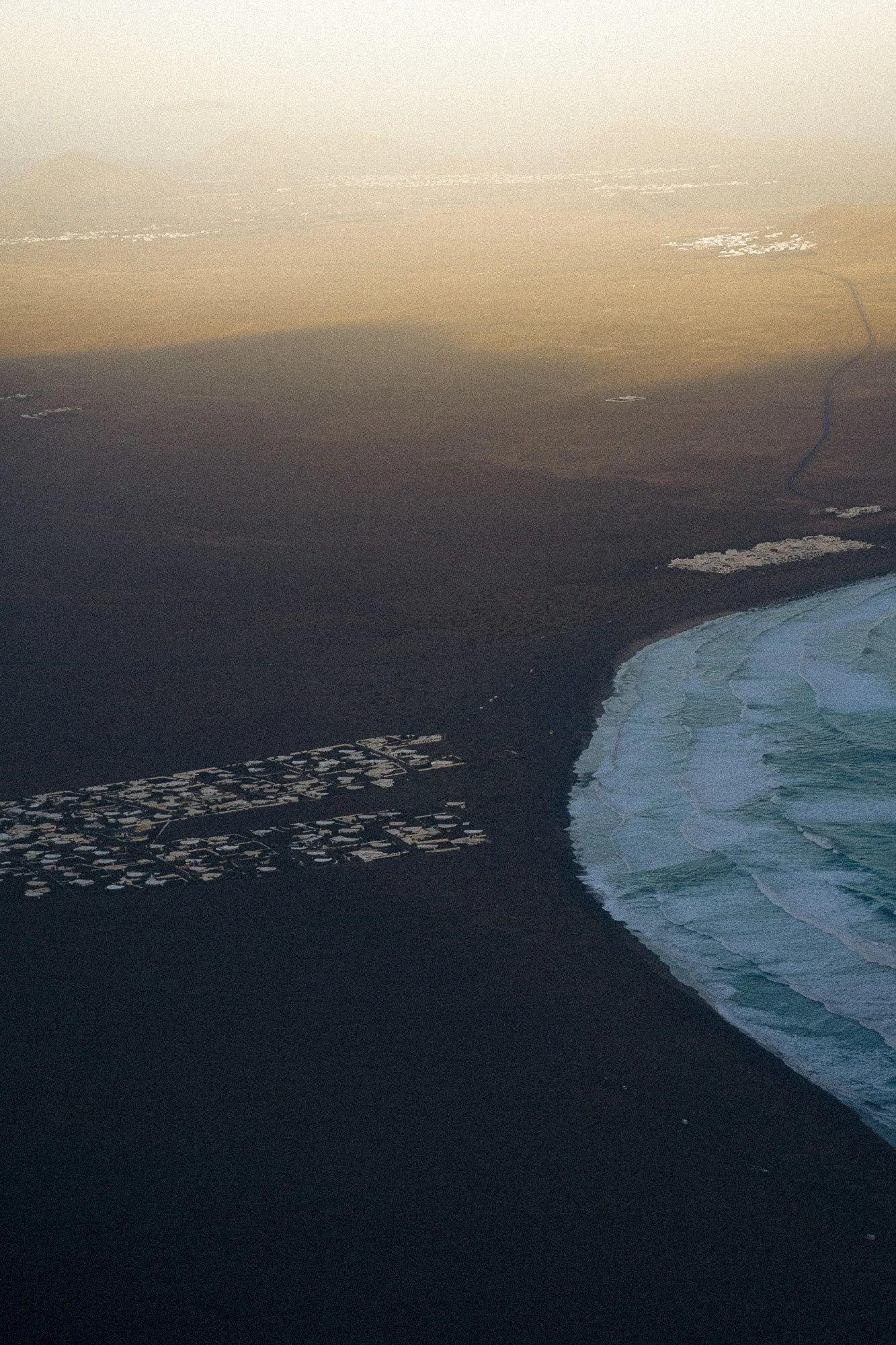 Aerial view of a coastal area with dark volcanic sand, white waves along the shoreline, and white structures or buildings on the land.