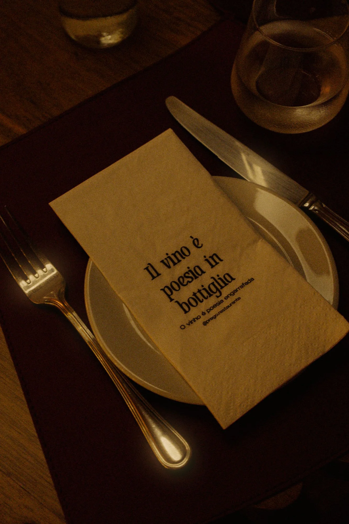 Una mesa con un plato blanco con una hoja de papel que tiene texto en italiano, un tenedor y un cuchillo, y dos vasos de agua, uno de los vasos se refleja en la superficie de la mesa de madera.