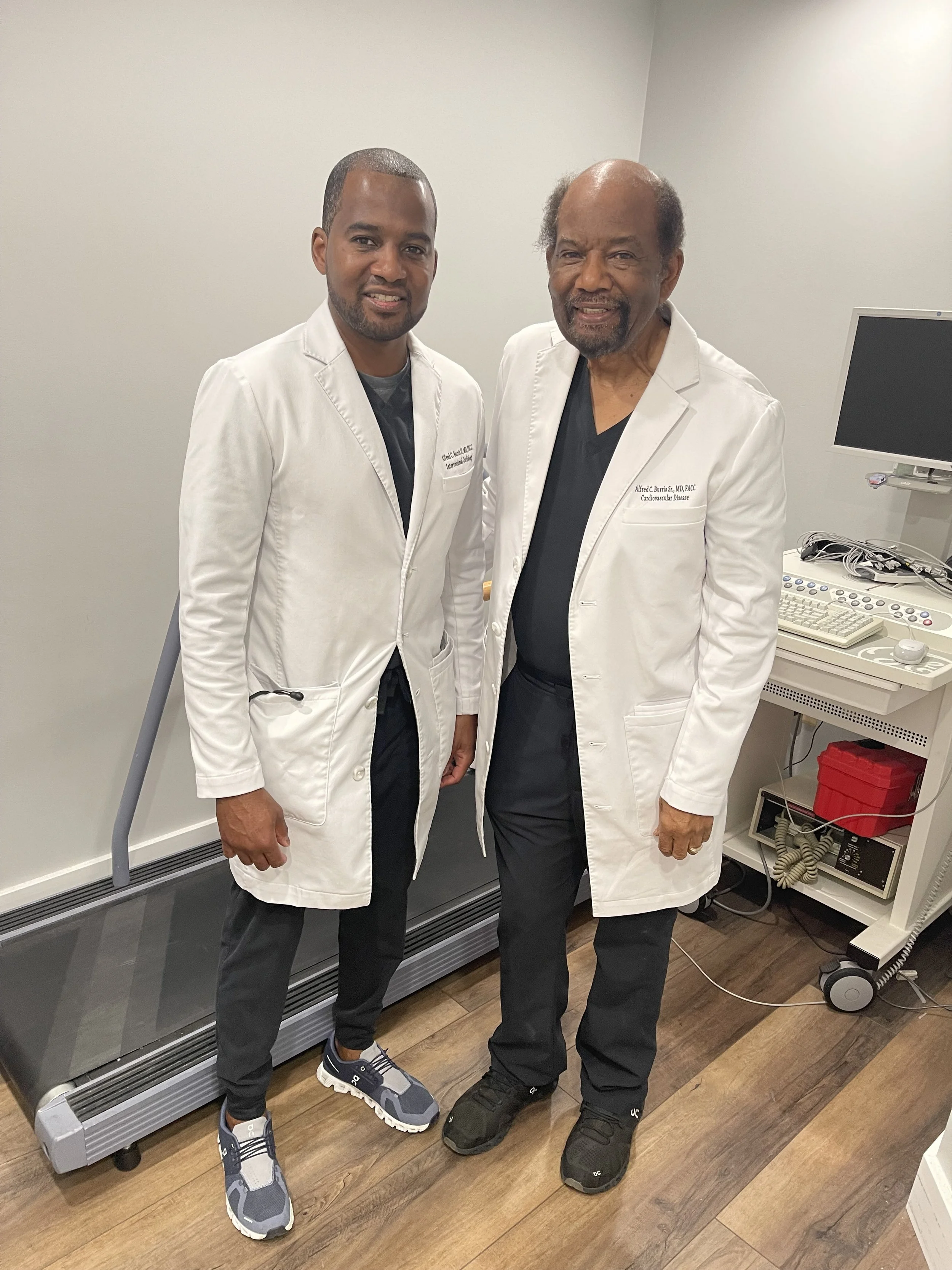 Two doctors in white lab coats standing in a medical room. They are smiling and posing for the photo.