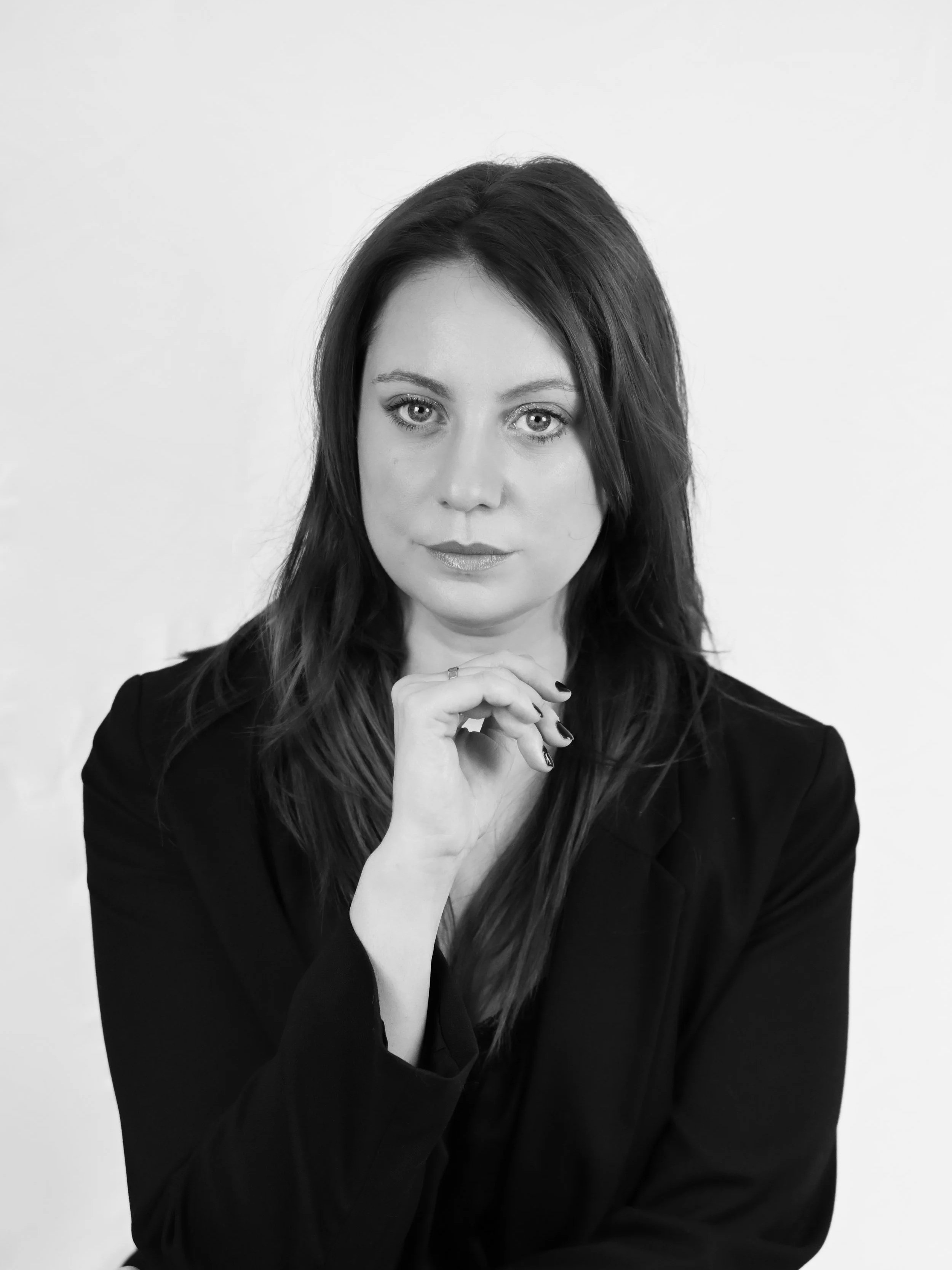 Portrait noir et blanc d'une femme avec les cheveux longs, regardant directement l'objectif, portant une veste noire, tenant sa main sous son menton, fond blanc.