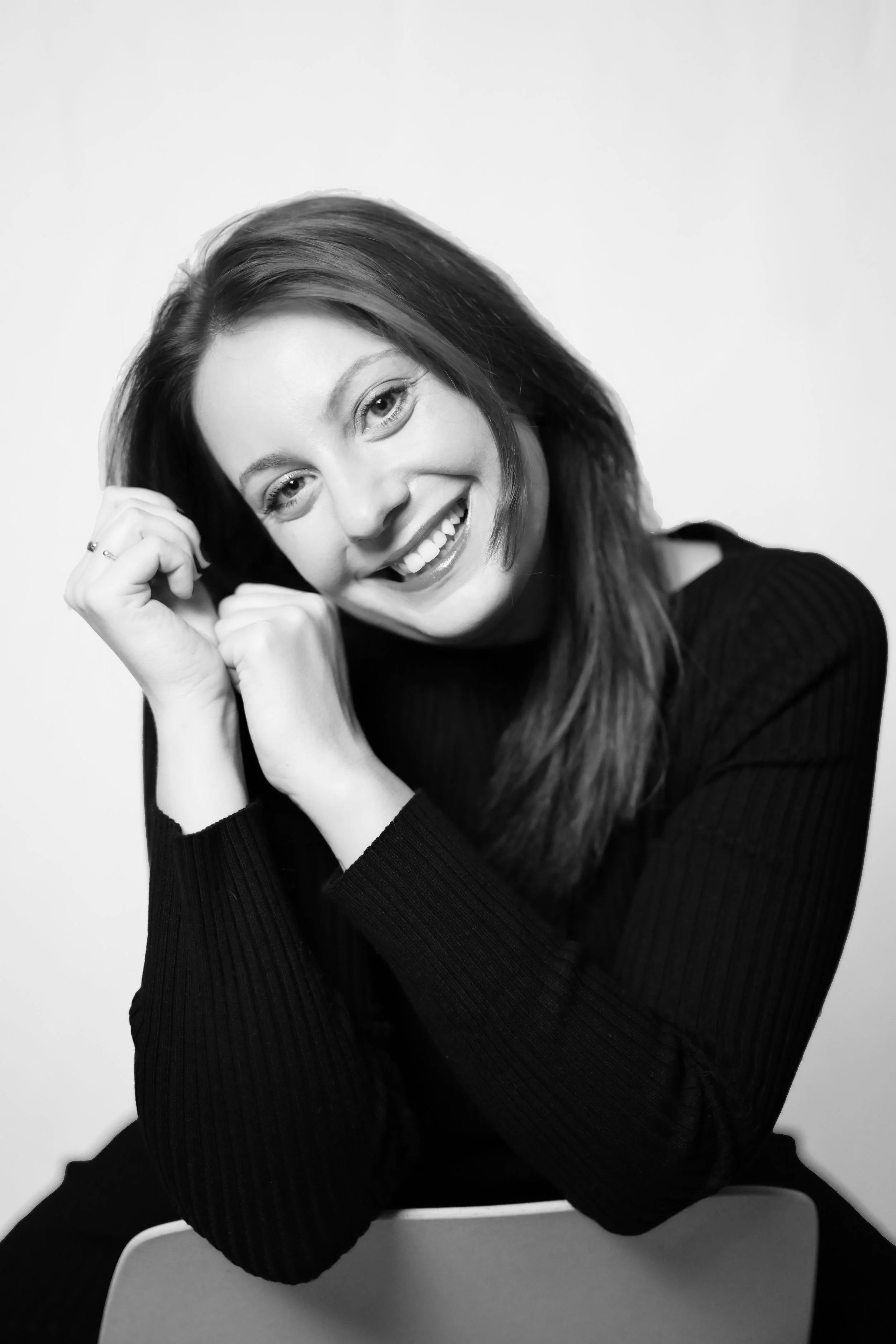 Portrait en noir et blanc d'une femme souriante, portant un pull noir, avec les cheveux bruns, assise près d'une chaise.