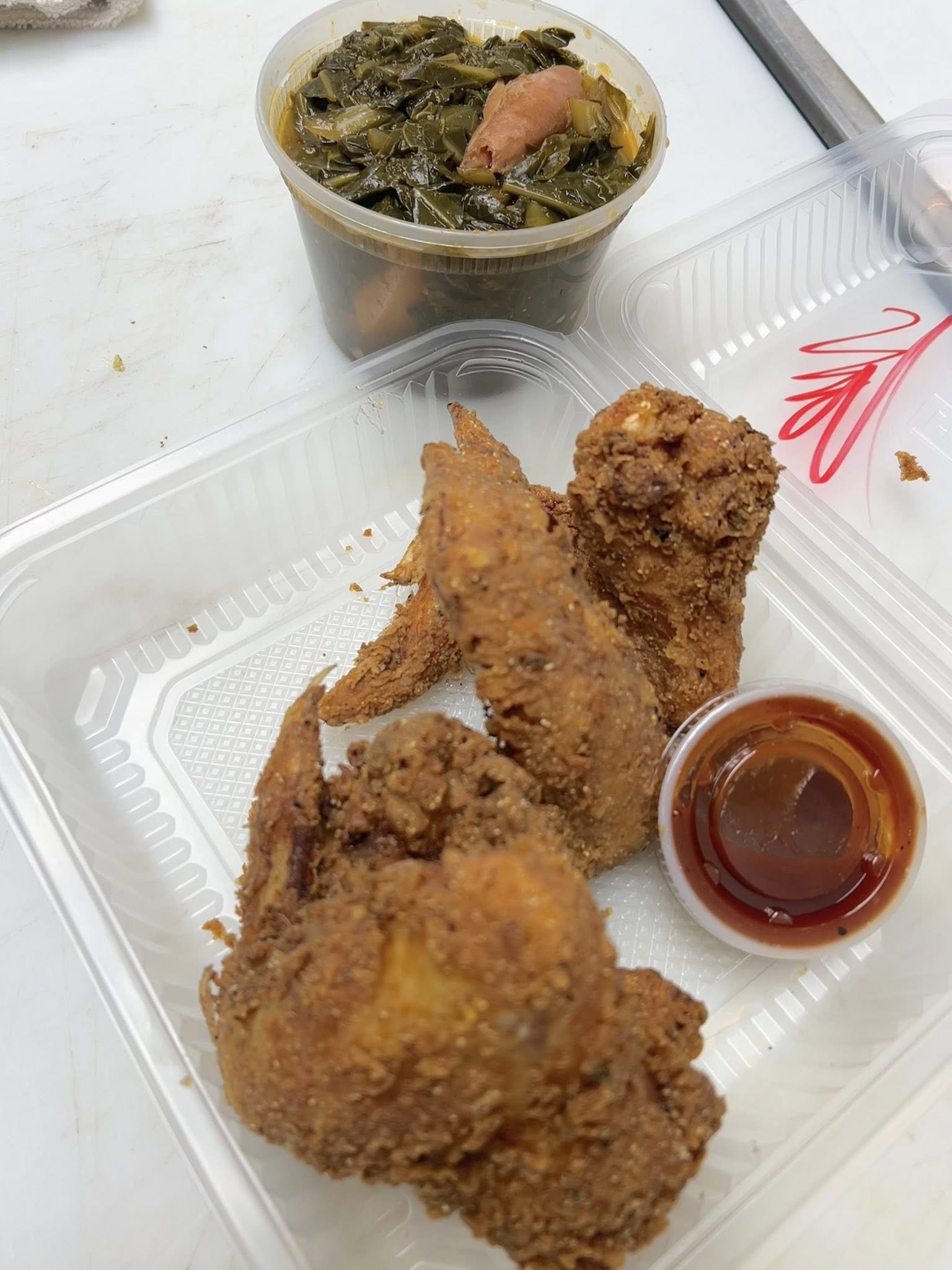 Fried chicken drumsticks with a side of dipping sauce in a takeout container and a cup of cooked collard greens with a ham hock.