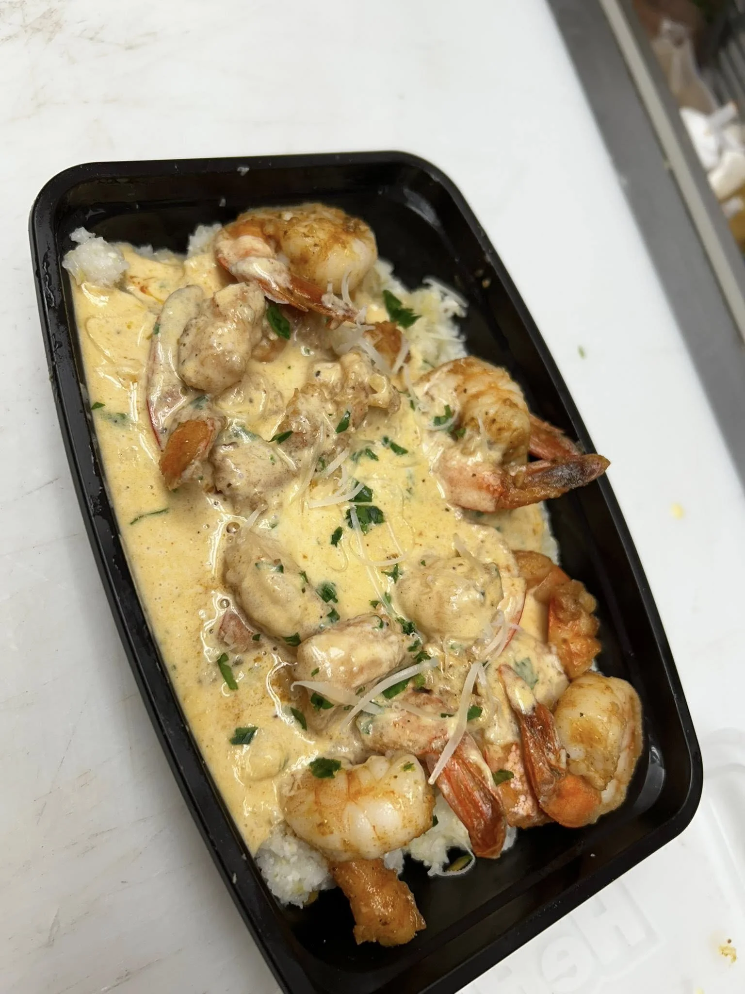 Shrimp and chicken in a creamy yellow sauce served over rice in a black container.