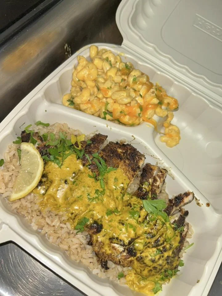 To-go container with rice, grilled meat topped with yellow sauce, lemon wedge, and chopped herbs; side of macaroni salad with carrots and herbs.