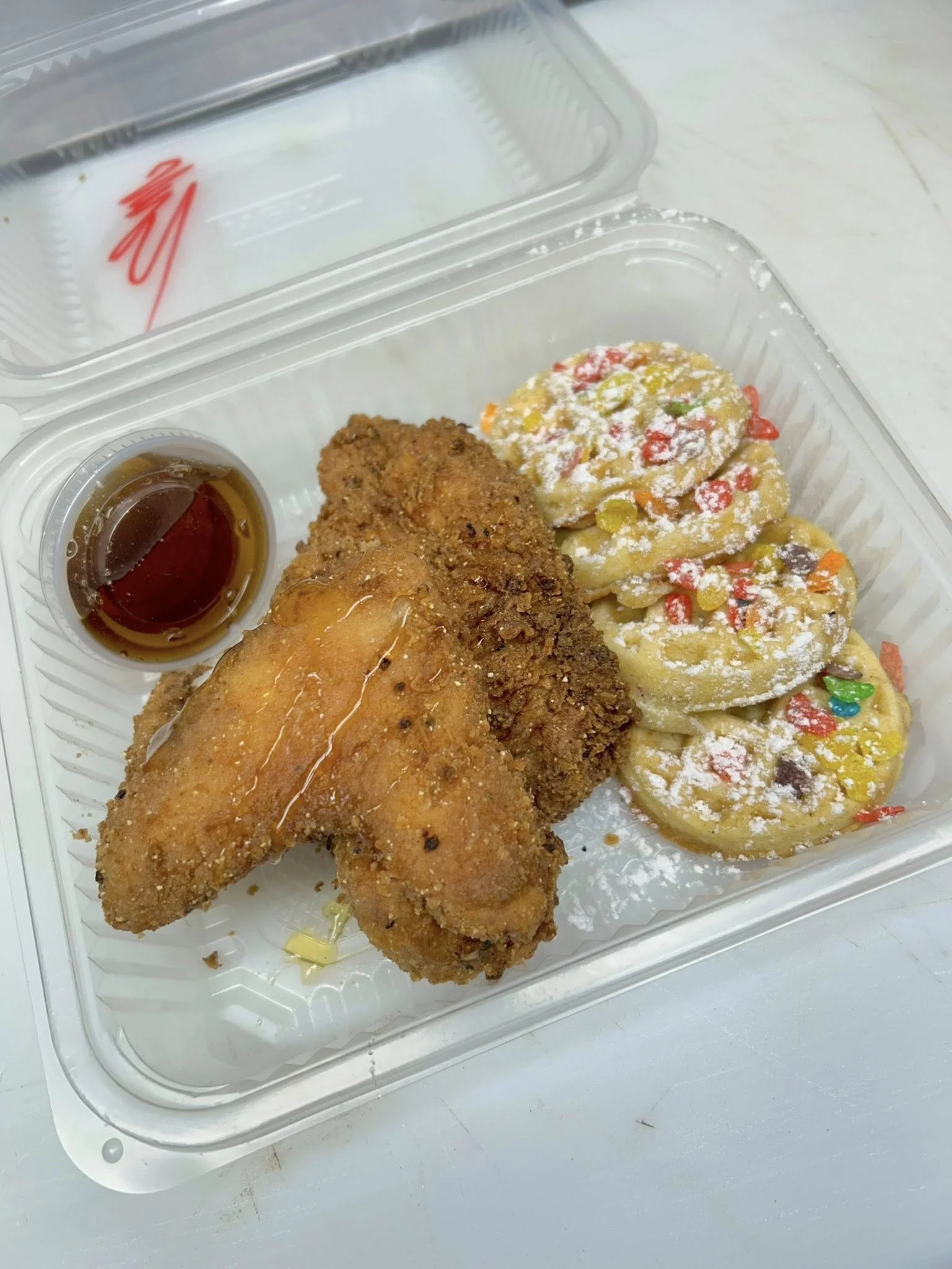 A takeout meal in a plastic container with fried chicken, two cookies with sprinkles and powdered sugar, and a small cup of syrup.