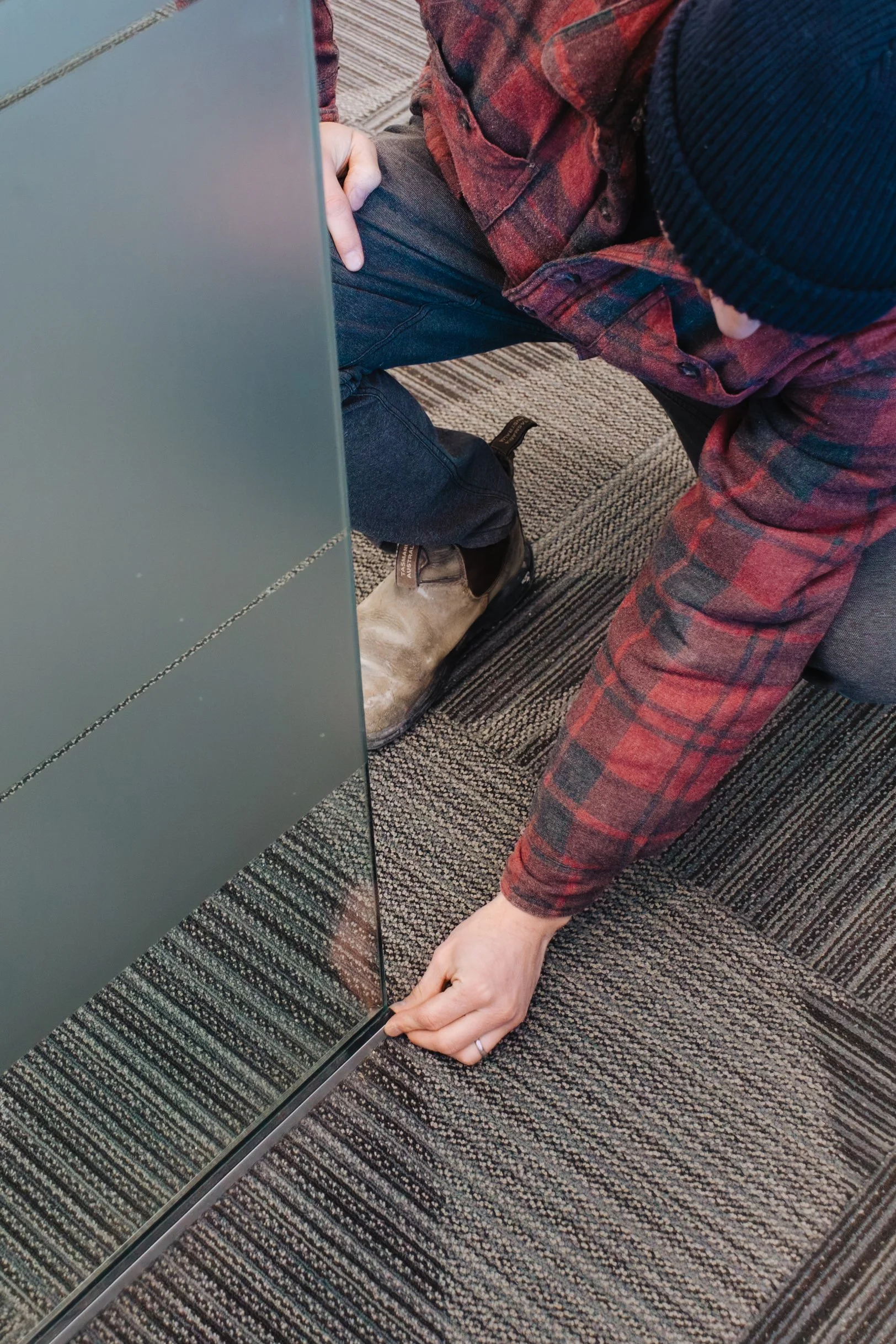 A person kneeling on the carpeted floor, installing or fixing a metal panel or door, wearing a dark blue beanie, red plaid shirt, and brown boots.