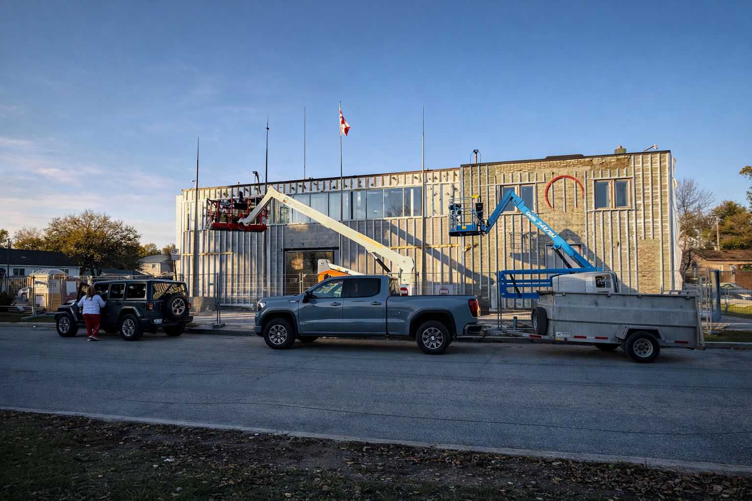 Construction workers installing exterior wall insulation on a building with two aerial work platforms, one red and one blue, parked along the street.