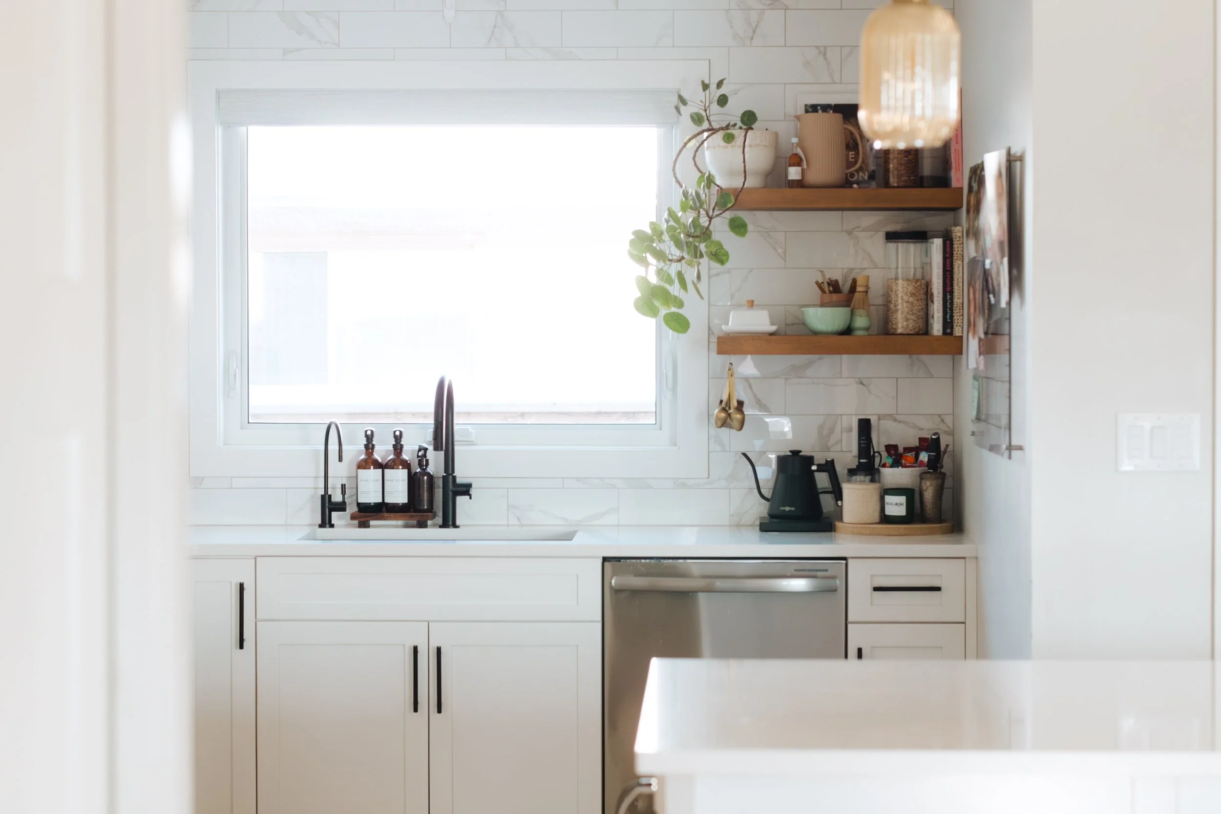 Modern kitchen with white cabinets, a large window above the sink, and wooden open shelves holding various kitchen items and plants.