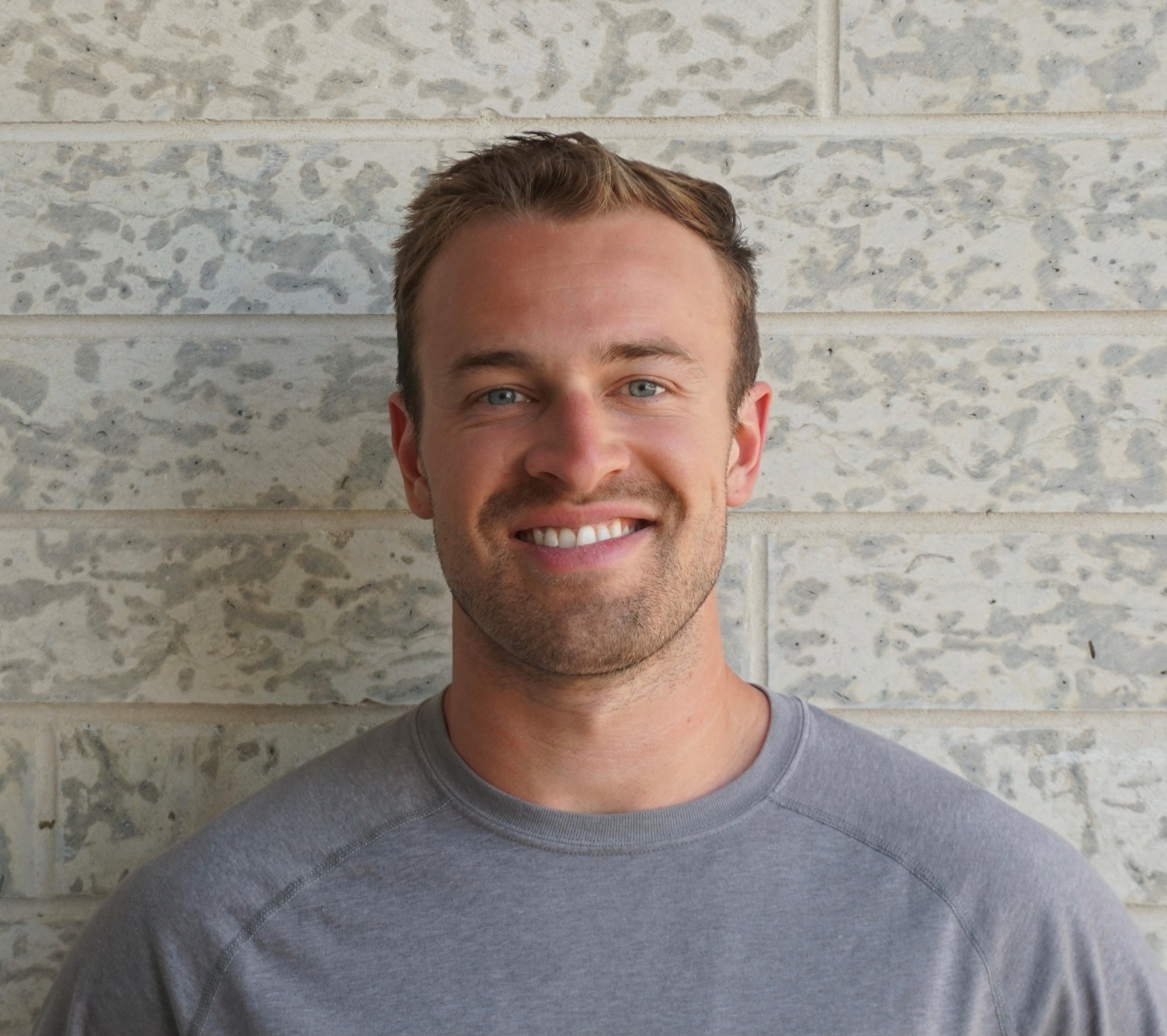 A young man with short, light brown hair and a beard, smiling, wearing a gray T-shirt, standing in front of a beige brick wall.