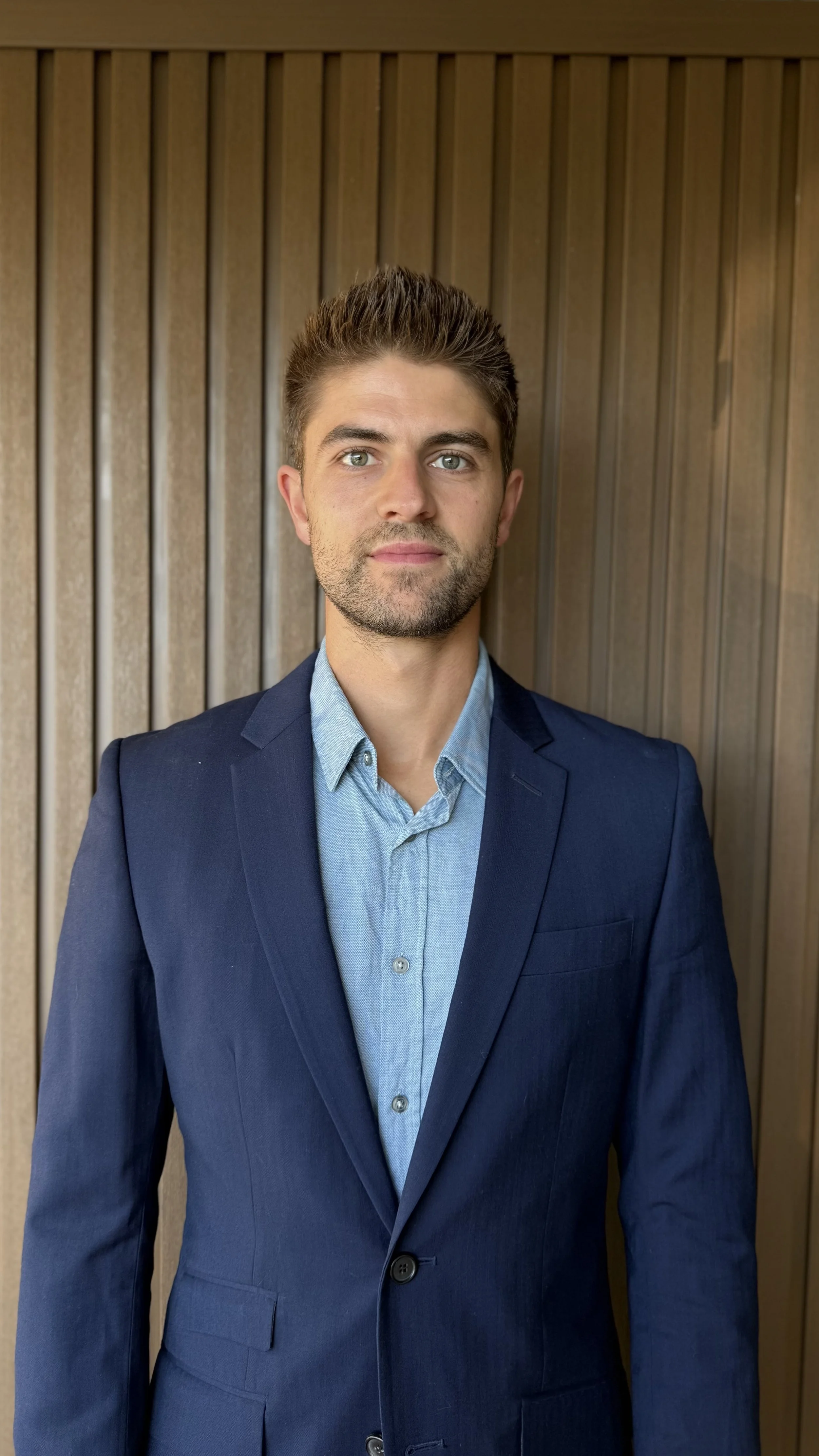 A smiling man with short light brown hair and blue eyes wearing a gray t-shirt standing in front of a textured stone wall.