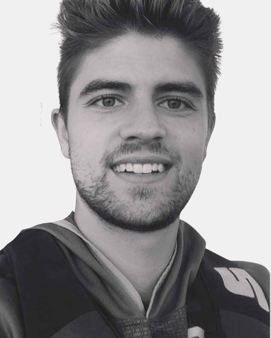 A smiling man with short light brown hair and blue eyes wearing a gray t-shirt standing in front of a textured stone wall.
