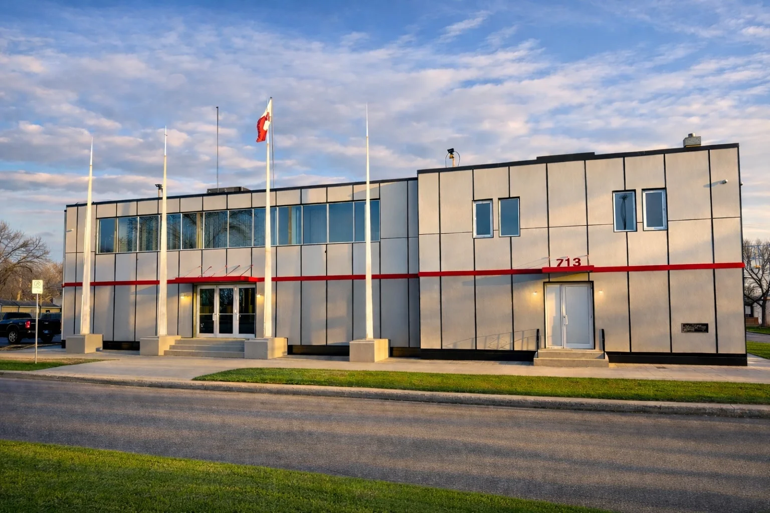 A modern two-story building with a white exterior and black lines, three flags on poles in front, and a red stripe with the number 713 above the door. Mirror windows on the upper floor and door stairs leading to the entrance.