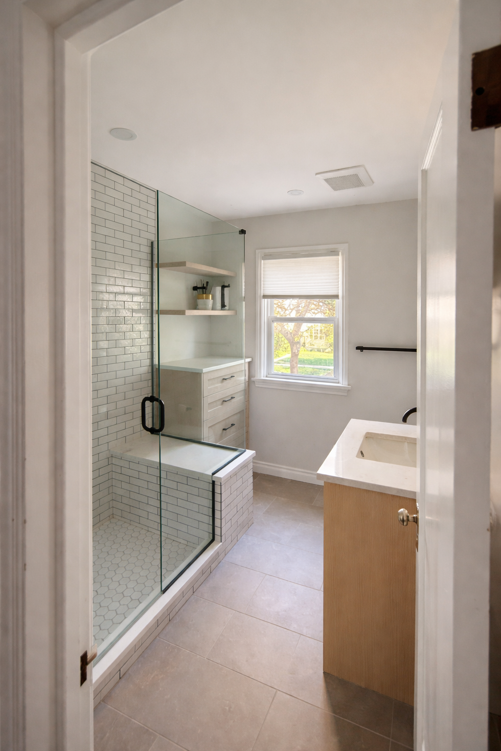 Bathroom with glass-enclosed shower, white subway tiles, built-in shelf and drawer unit, window with blinds, and sink with wooden cabinet.