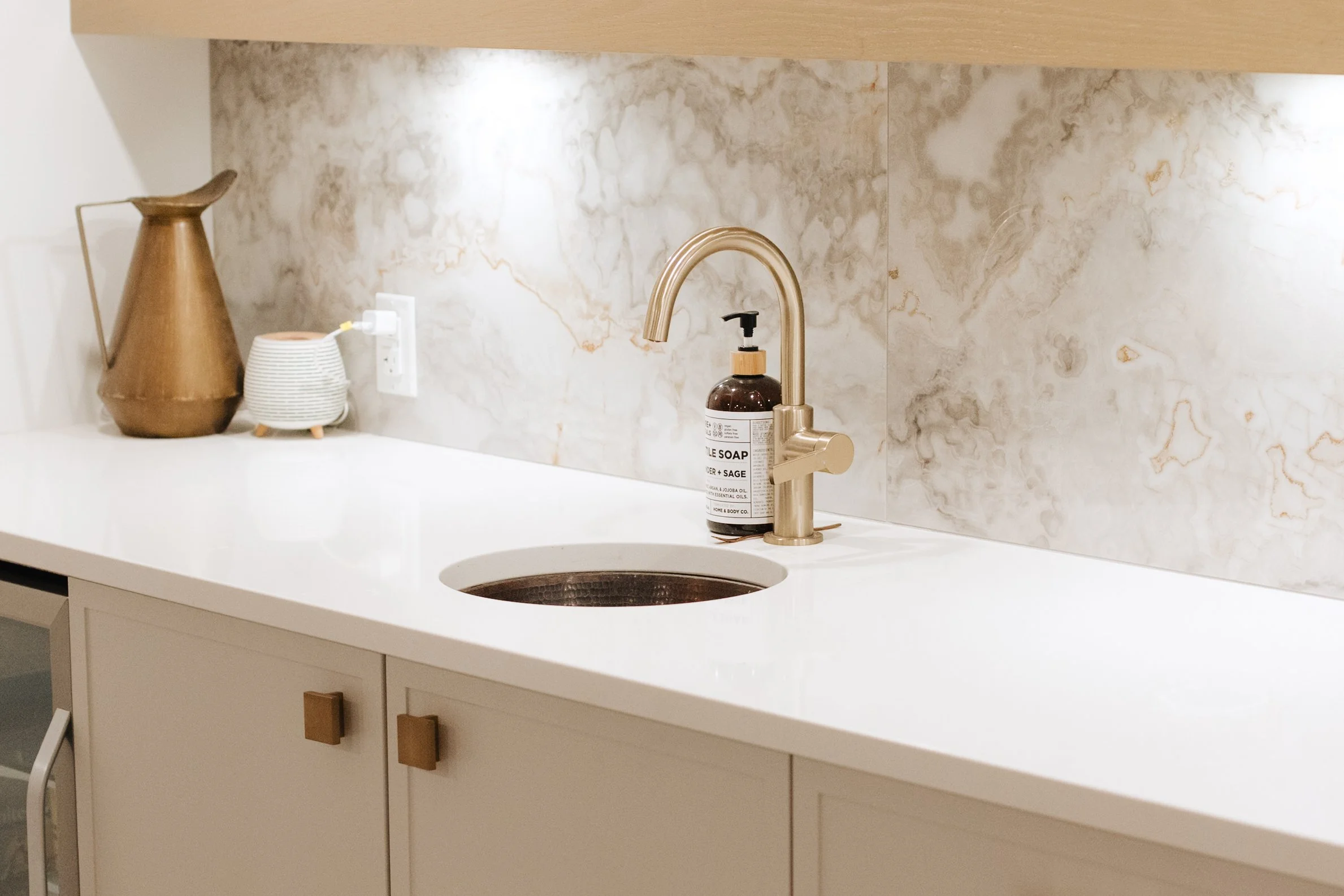 White bathroom countertop with a beige marble tile backsplash, a gold faucet, a soap dispenser, a brown pitcher, a small bowl, and a cabinet with square handles.