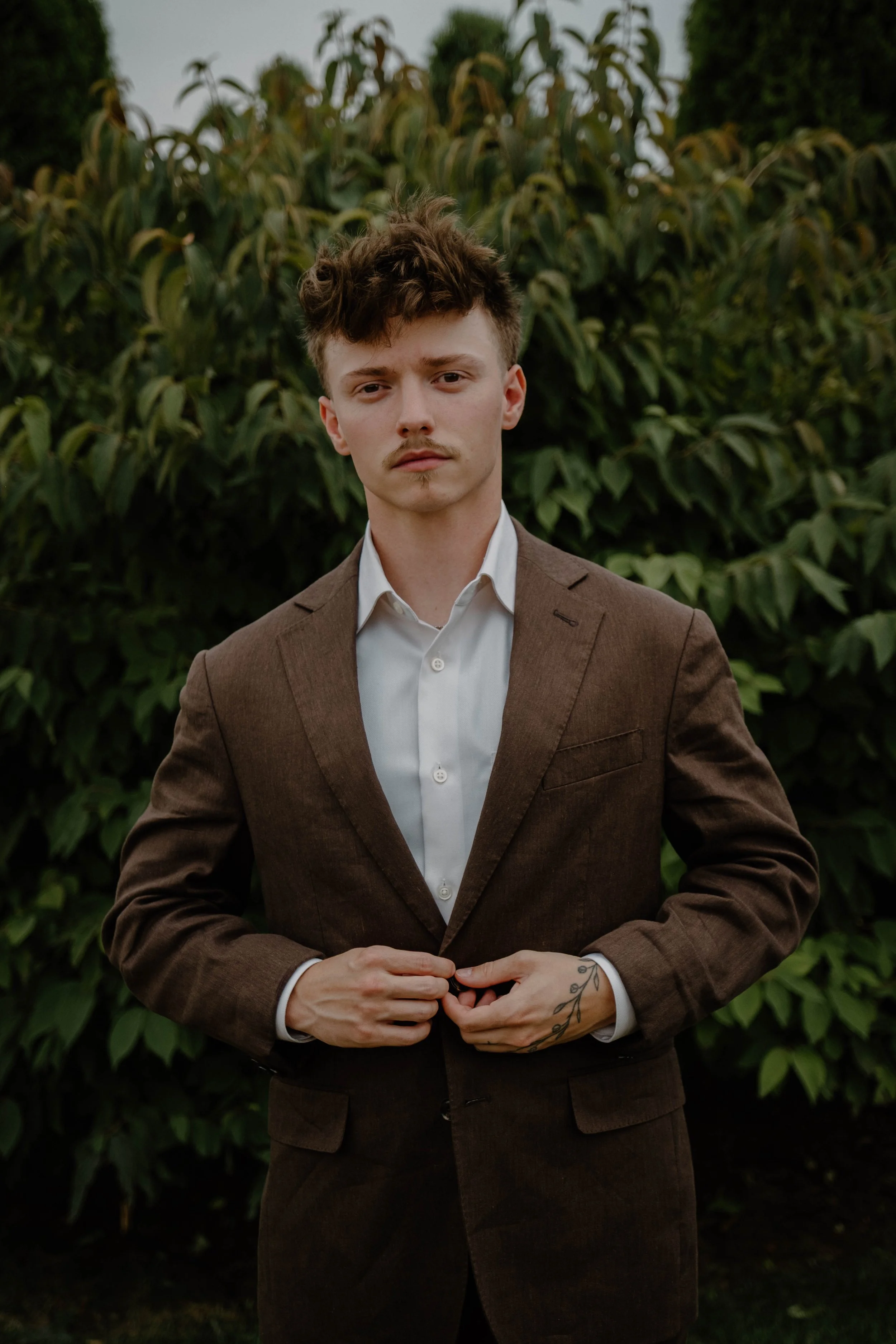 A young man with light skin and wavy brown hair, wearing a white dress shirt and brown blazer, standing in front of green foliage, adjusting his blazer with a neutral expression.