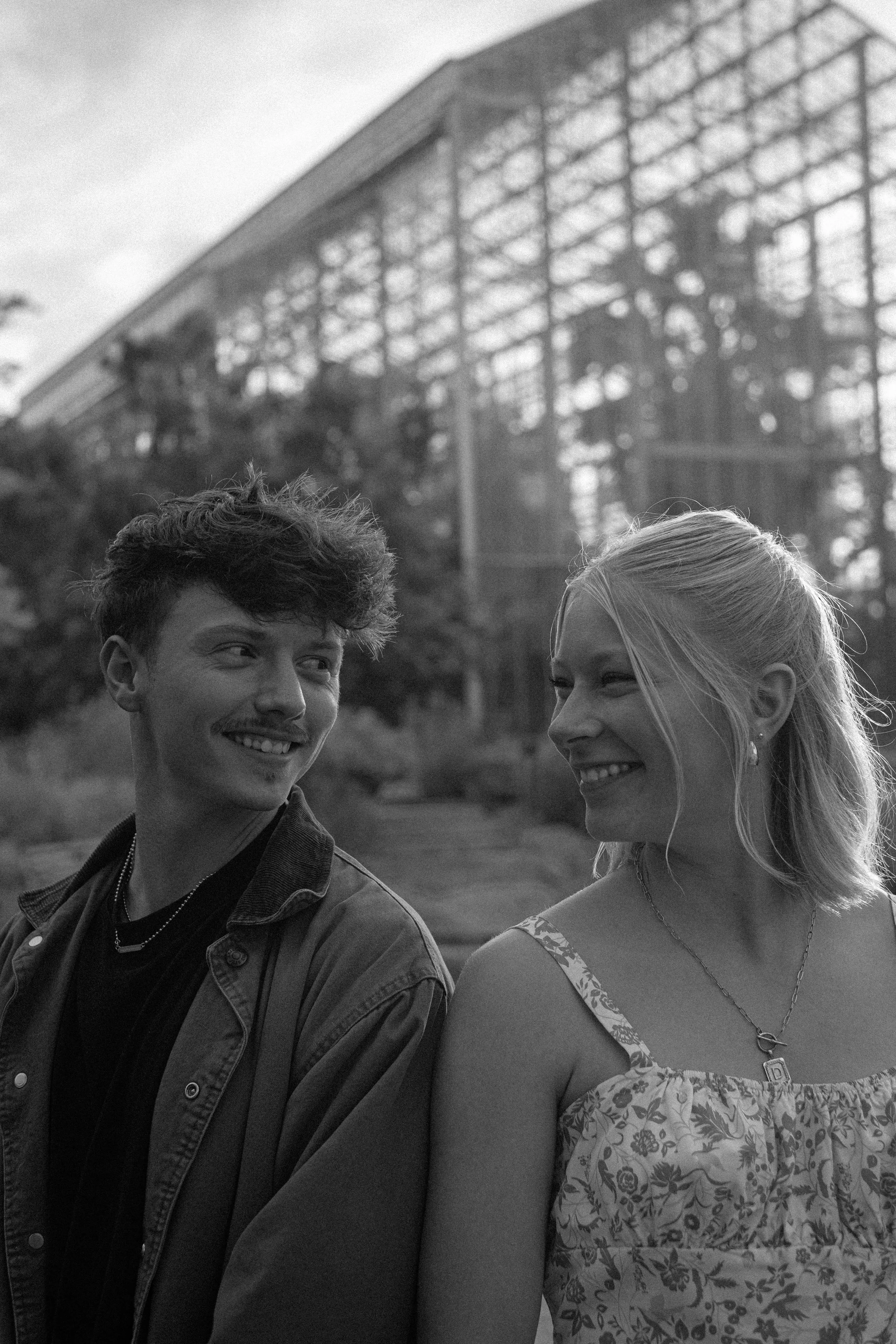 A black and white photo of a smiling young man with curly hair and a young woman with straight hair in a floral dress, standing outdoors with a building in the background.