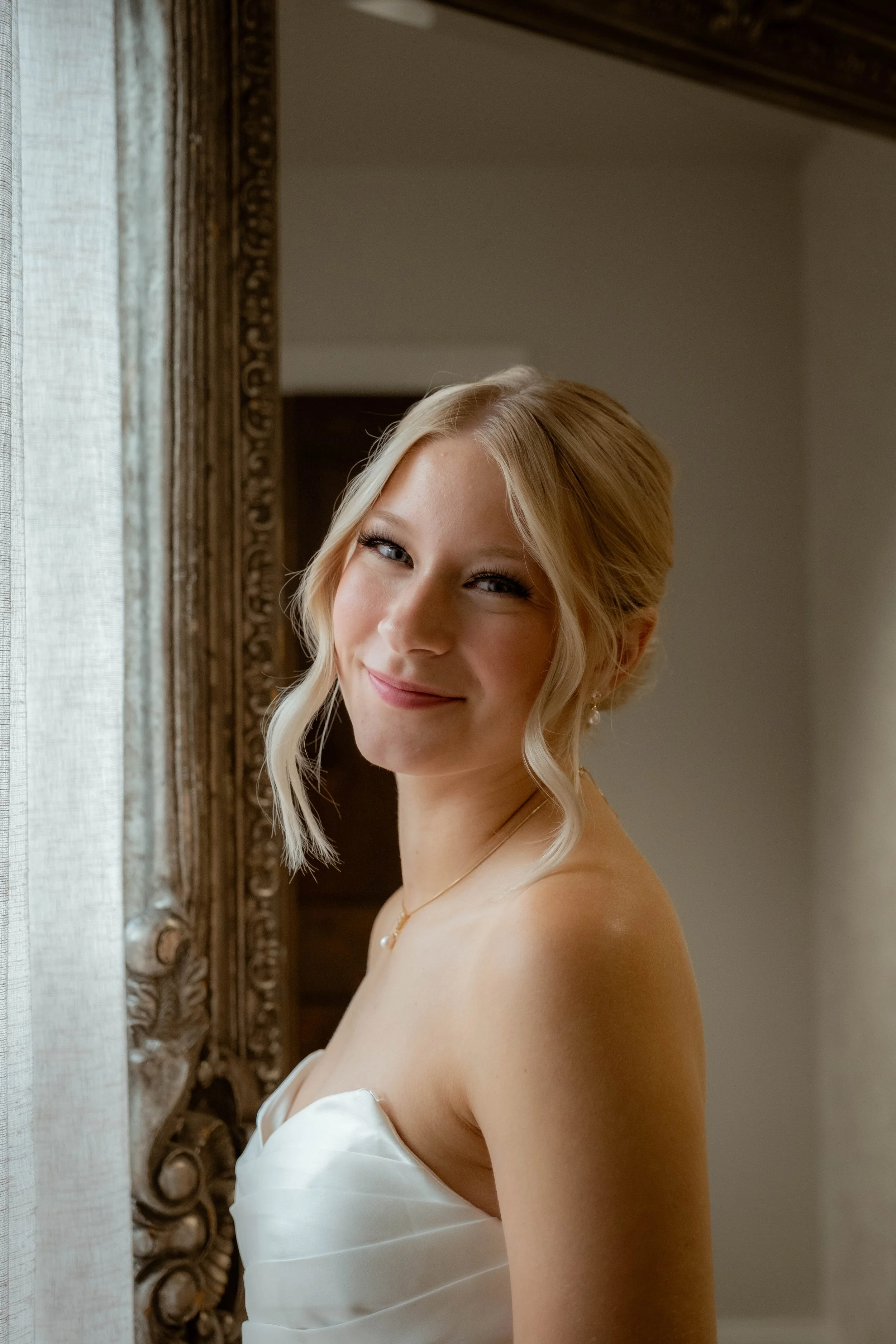 A young woman with blonde hair styled in loose waves, wearing a strapless white dress, standing by a window with a sheer curtain and ornate gold-framed mirror, smiling softly at the camera.