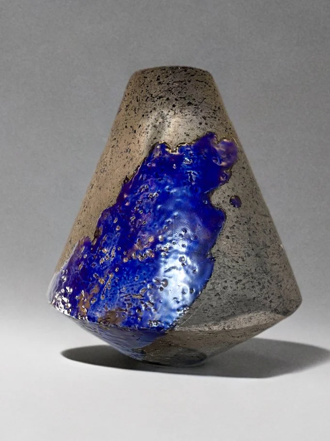 A modern, cone-shaped sculpture made of concrete with a blue glass or resin accent on one side, casting a shadow on a gray surface.