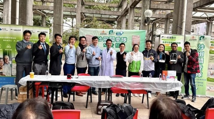 Group of people standing behind a table at an outdoor event or presentation, with banners and promotional materials in the background.