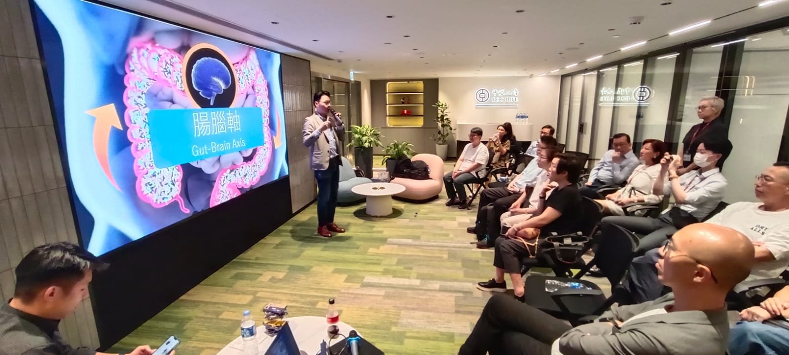 A presenter standing in front of a large screen showing a diagram of the gut-brain axis, speaking to an audience seated in a modern conference room. The audience includes men and women, some taking notes or photos, with several wearing face masks.