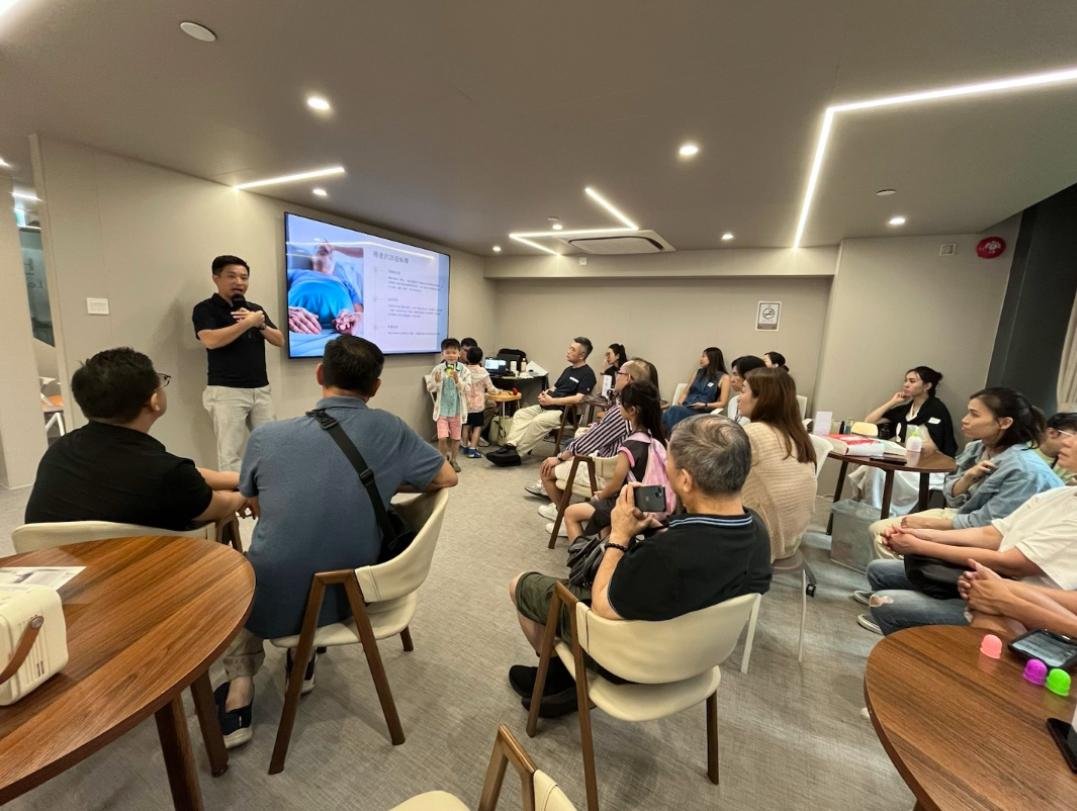 People attending a presentation or meeting in a modern conference room, with a speaker at the front and a large screen displaying a slide.