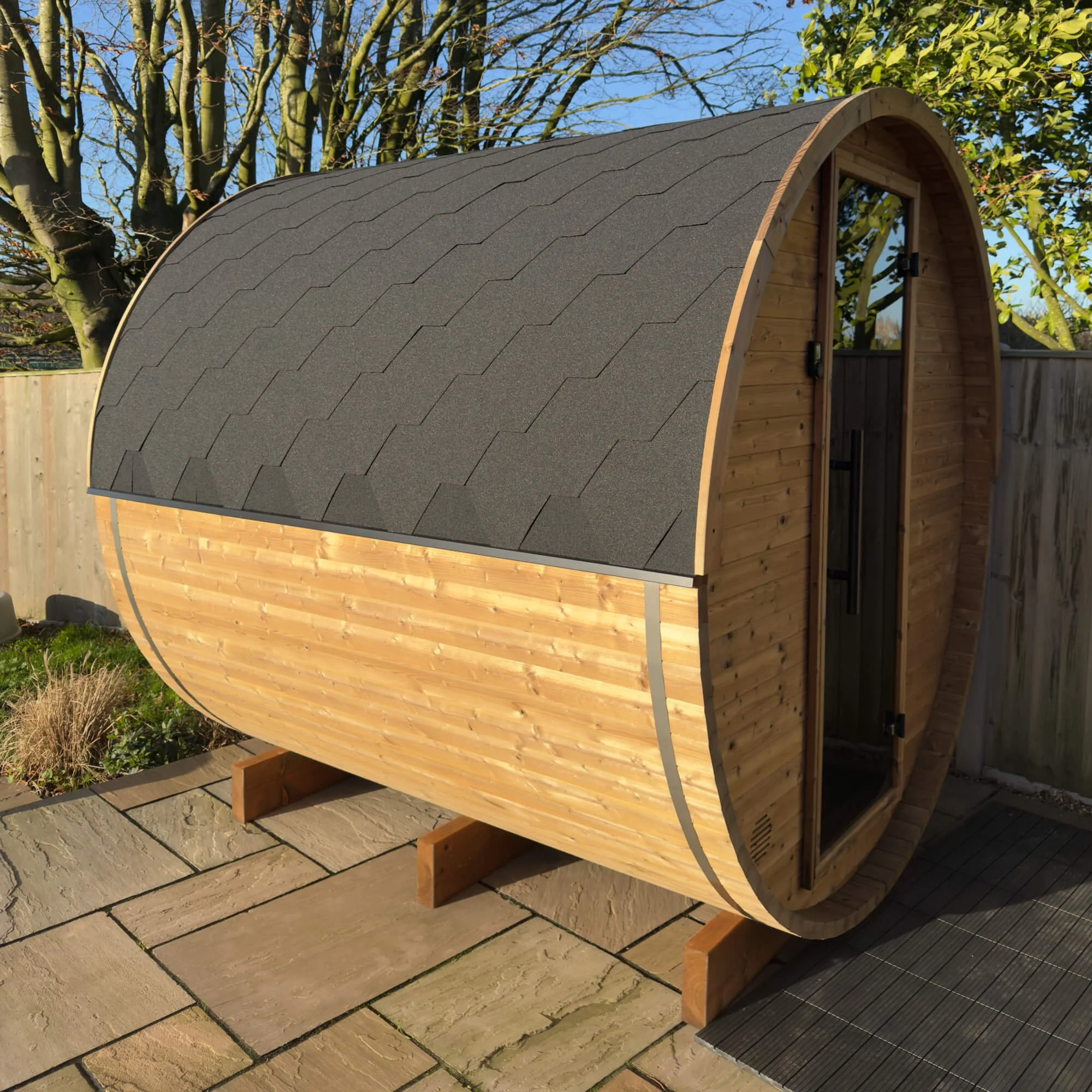 A small wooden outdoor sauna with a curved, black shingled roof and a narrow door, situated on a tiled patio in a backyard with trees and a wooden fence.