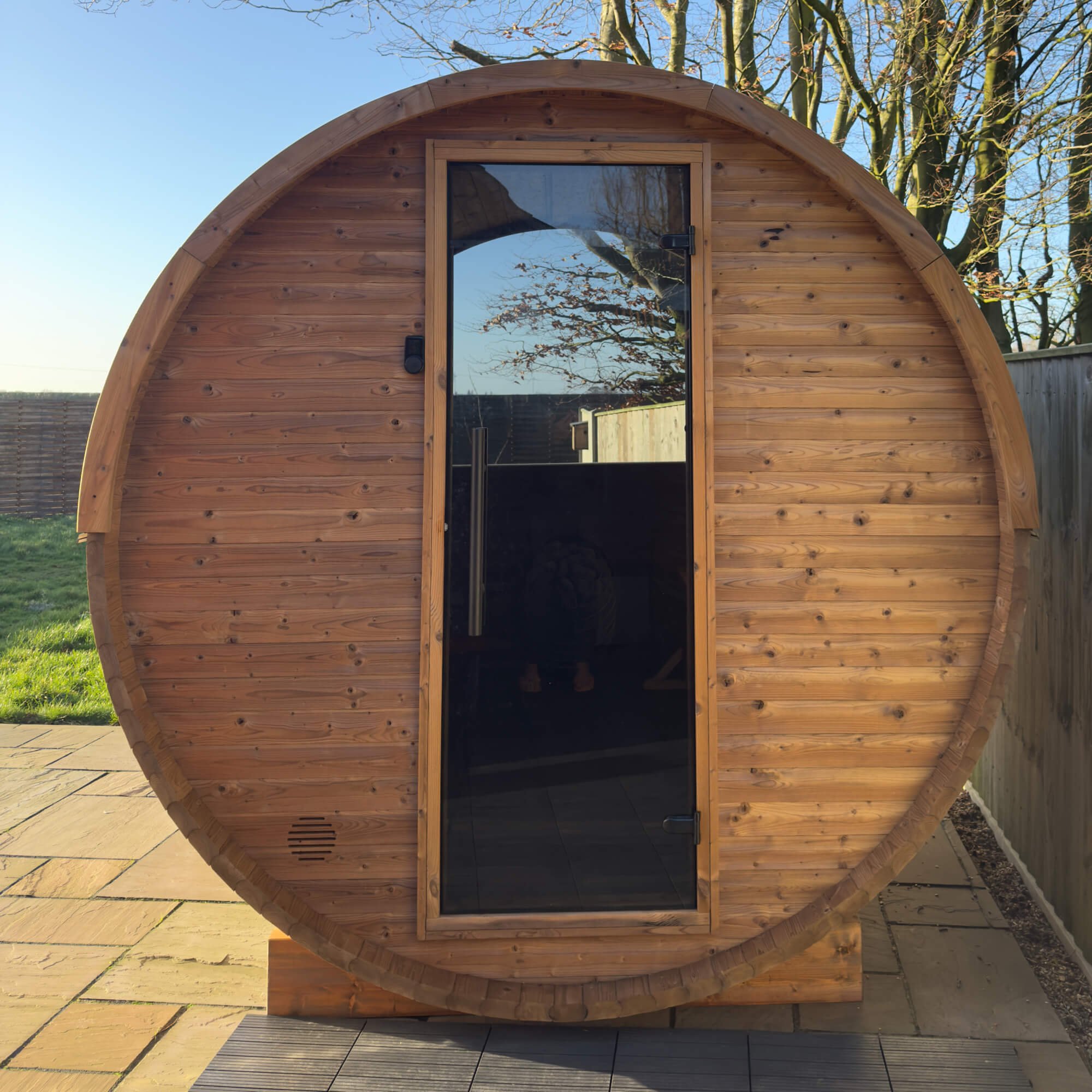 A round wooden sauna with a glass door, situated outdoors on a tiled patio, with a blue sky and trees in the background.