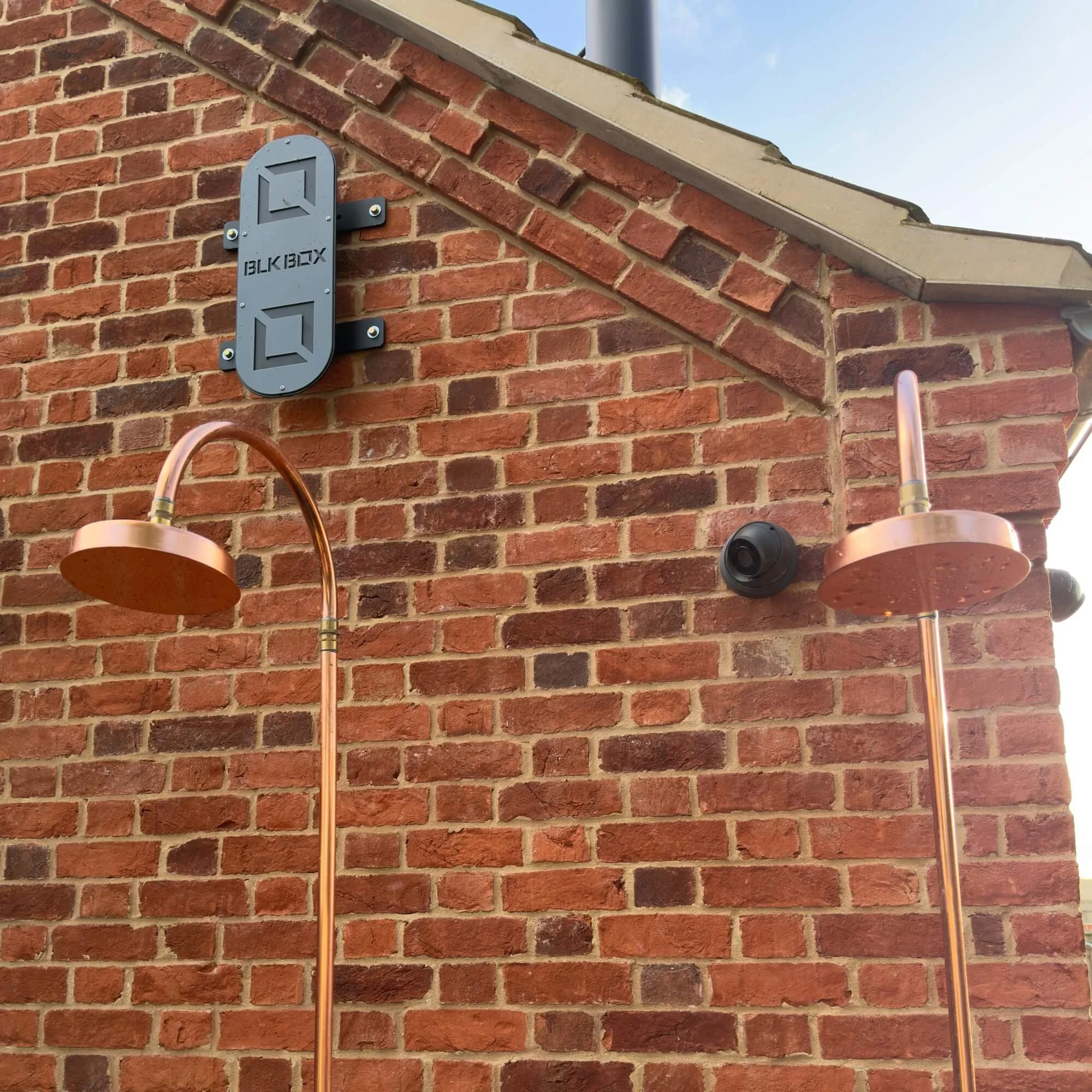 Close-up of a brick wall exterior with a black security camera and two copper outdoor shower heads. A black BBK Box mounted on the wall and part of a building roof and sky are visible.