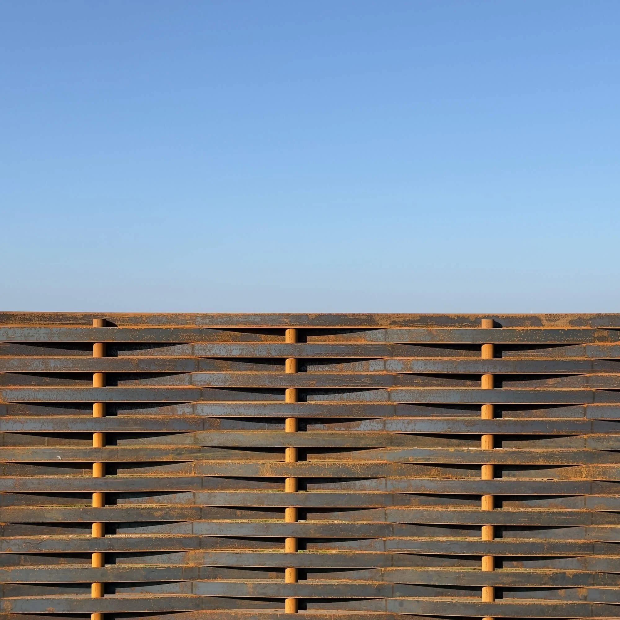 Close-up of a rusty metal structure with horizontal slats and cylindrical support rods, with a clear blue sky in the background.