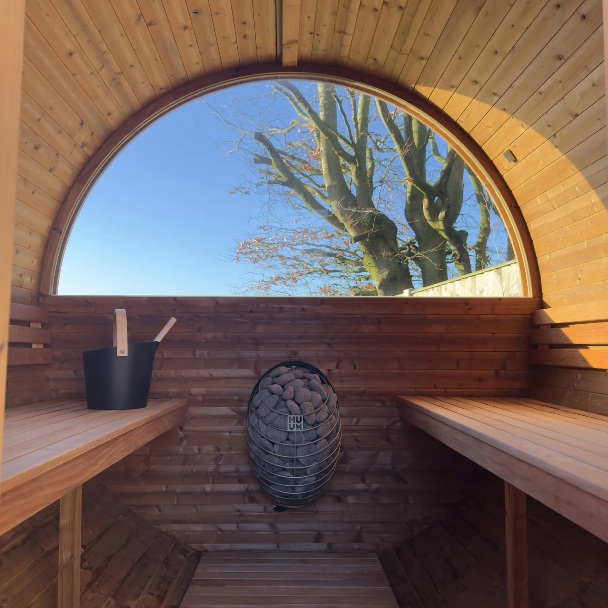 Interior of a wood-paneled sauna with a large arched window showing a tree outside and a clear blue sky, sauna heater with stones, and wooden benches.