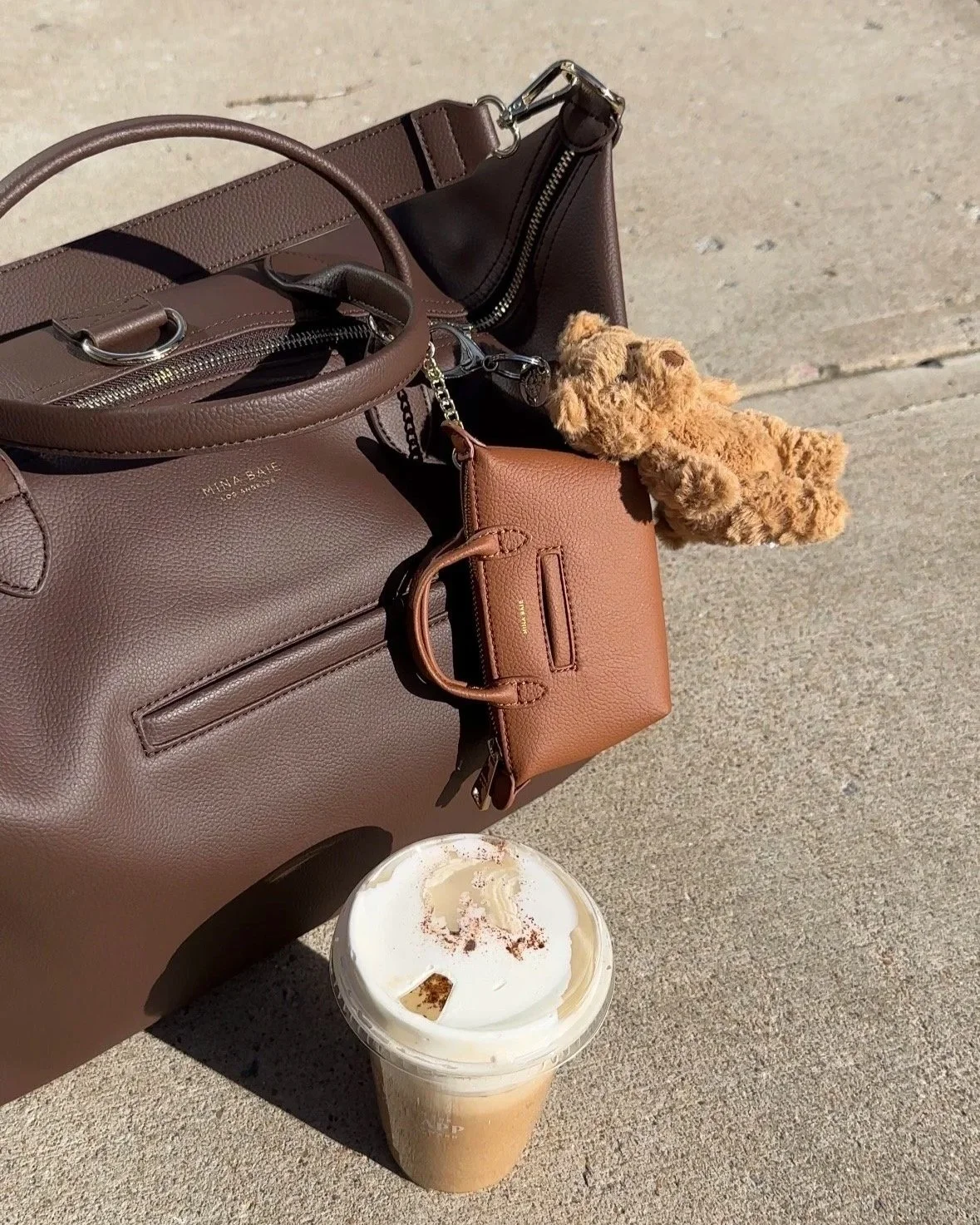 Multiple handbags in shades of brown placed on concrete ground with a small plush teddy bear hanging from one of the bags, and a coffee drink with whipped cream on top in a plastic cup nearby.