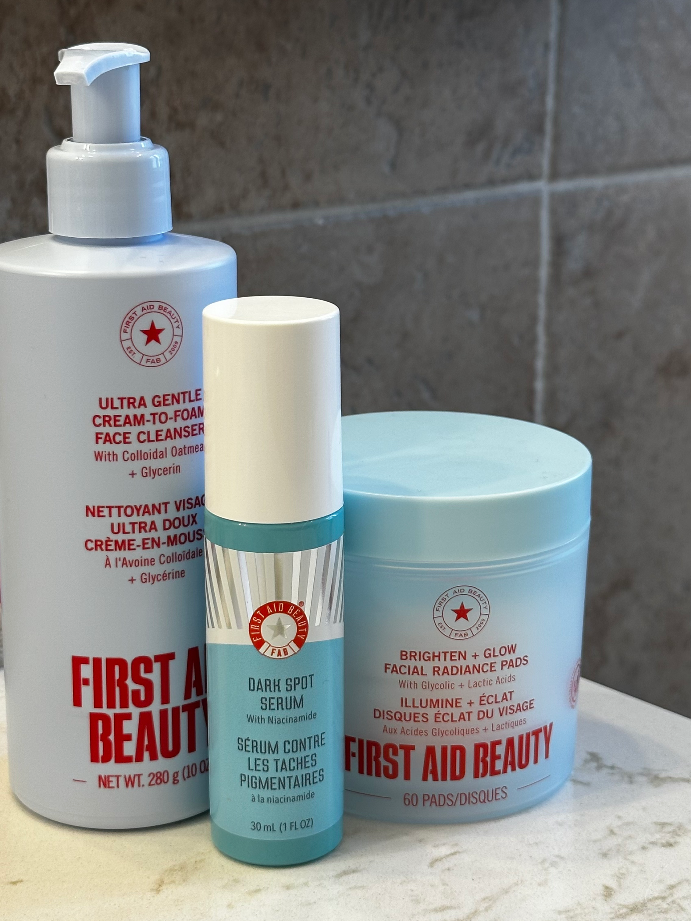 A collection of skincare products, including a bottle of face cleanser, a bottle of dark spot serum, and a container of facial radiance pads, all labeled with the First Aid Beauty brand.