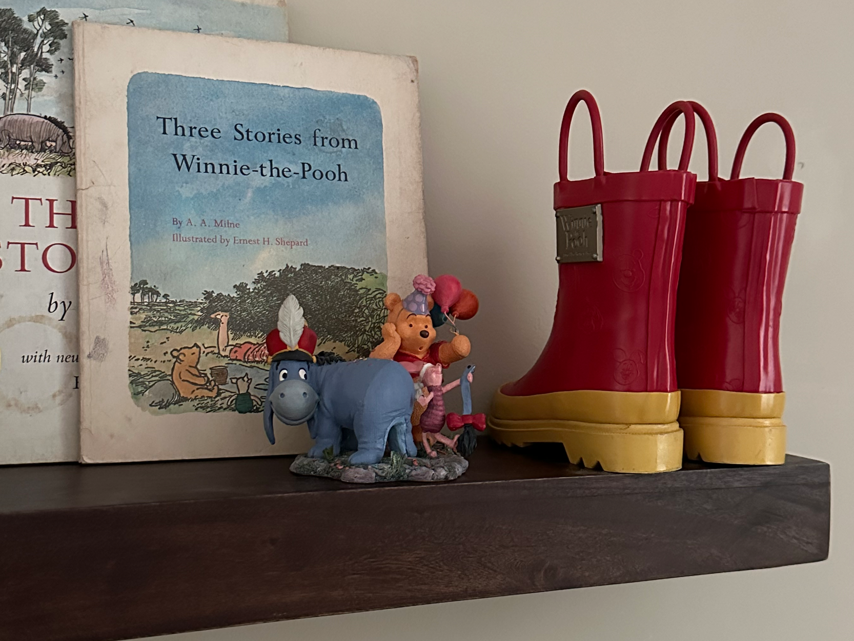 A shelf displays a children's book titled 'Three Stories from Winnie-the-Pooh,' two Winnie-the-Pooh figurines, and a pair of red and yellow rain boots.